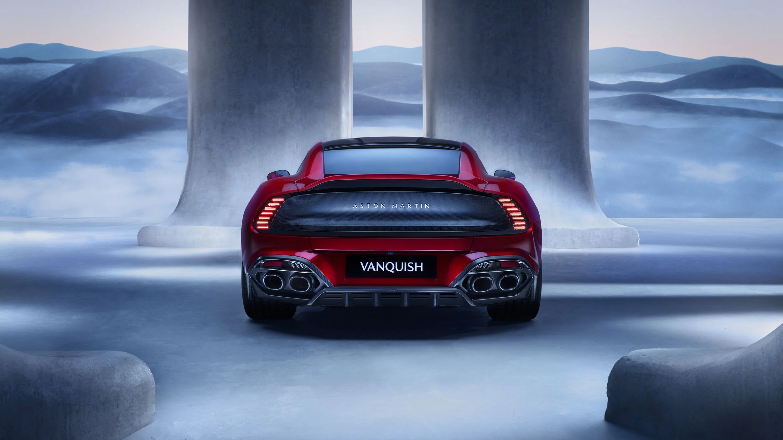 Rear view of a red Aston Martin Vanquish sports car parked outdoors with large stone columns in the background and mountainous scenery in the distance.