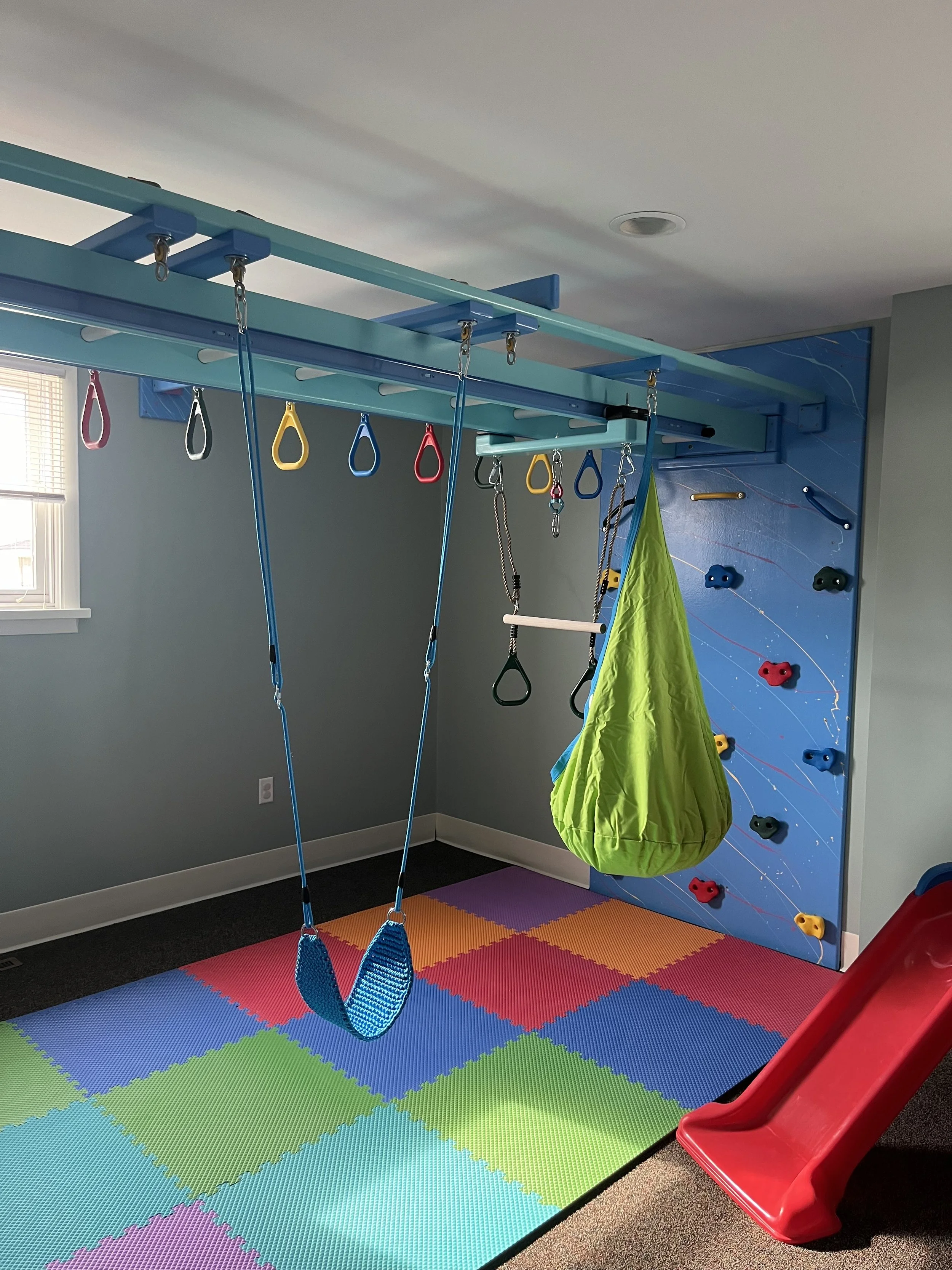 Indoor children's sensory gym area with climbing wall, yellow, red, and blue handles, swings, and a red slide, set on a colorful foam mat.