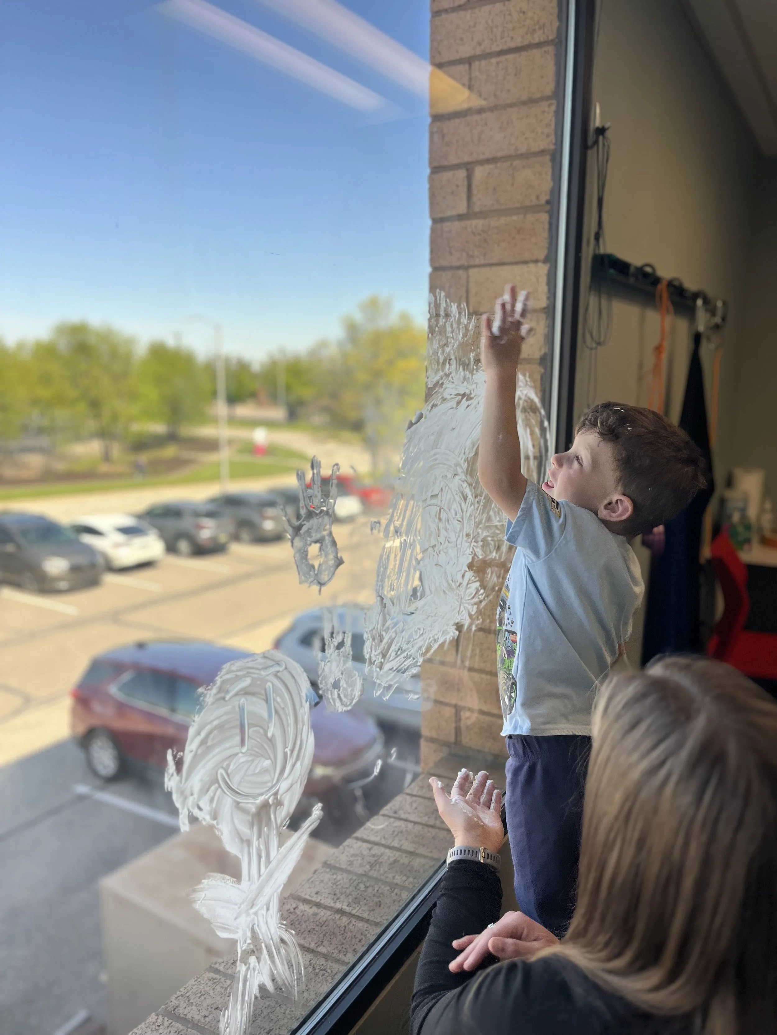 A young boy is engaging in sensory play with his occupational therapist by smearing shaving cream on a large glass window. The outside view shows a parking lot with cars, trees, and a clear blue sky.