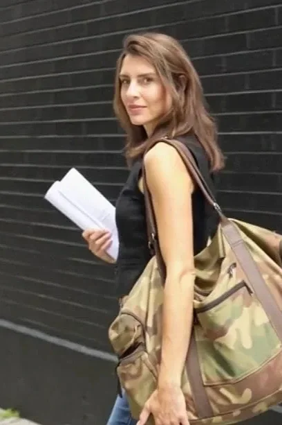 A young woman with shoulder-length brown hair stands outdoors near a black brick wall, holding a notebook or folder in her right hand, wearing a sleeveless black top, with a large camouflage shoulder bag on her left shoulder.