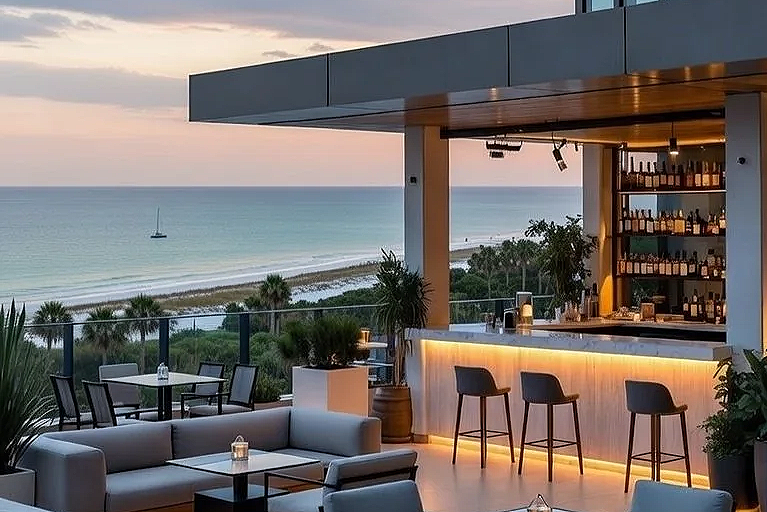 Outdoor bar and seating area overlooking the ocean during sunset, with a sailboat in the water.
