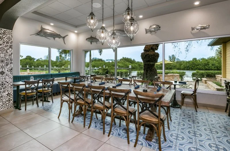 Interior of a restaurant with large windows overlooking a river, featuring wooden dining tables and chairs, decorative fish wall art, and hanging light fixtures.