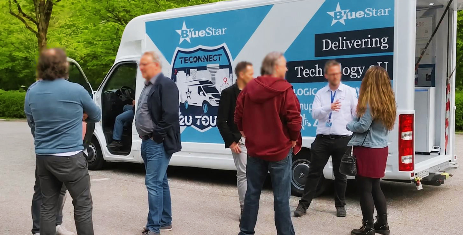 Grupo de personas conversando alrededor de un camión de entrega con logotipo y texto sobre tecnología y transporte, en un área exterior rodeada de árboles verdes.
