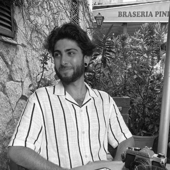 Un joven con barba y cabello oscuro, usando una camisa de rayas, se encuentra sentado en un lugar con plantas y un muro de piedra al fondo, en un entorno al aire libre.