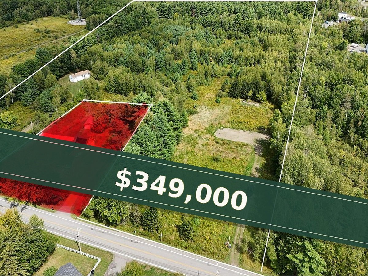 Aerial view of a large, mostly wooded plot of land outlined with white borders. Part of the land is highlighted in red, showing structures and trees, with a road running along the front edge and a small car parked on a driveway.