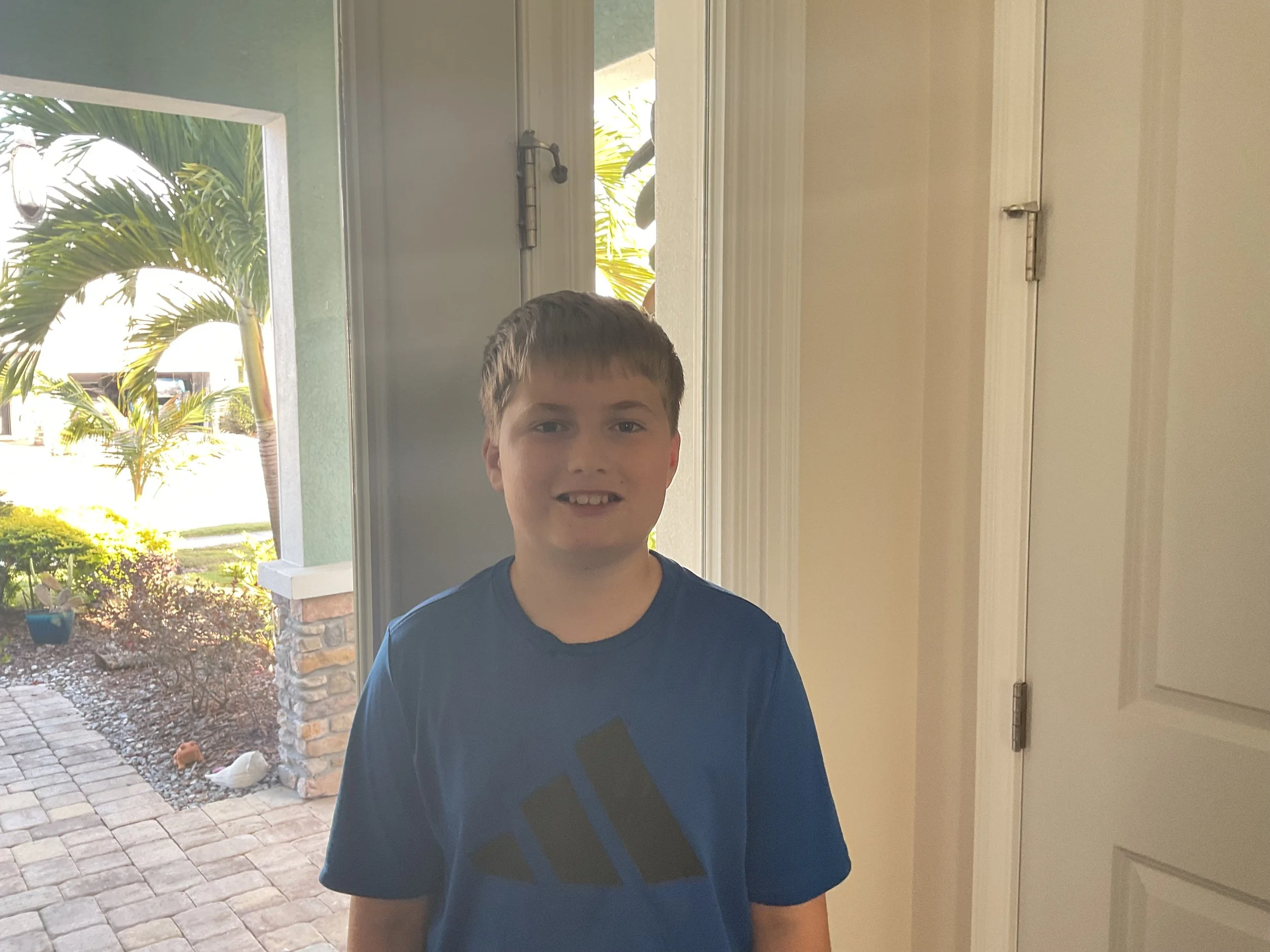 Close-up of a young boy with brown hair and big eyes, smiling against a plain wall.