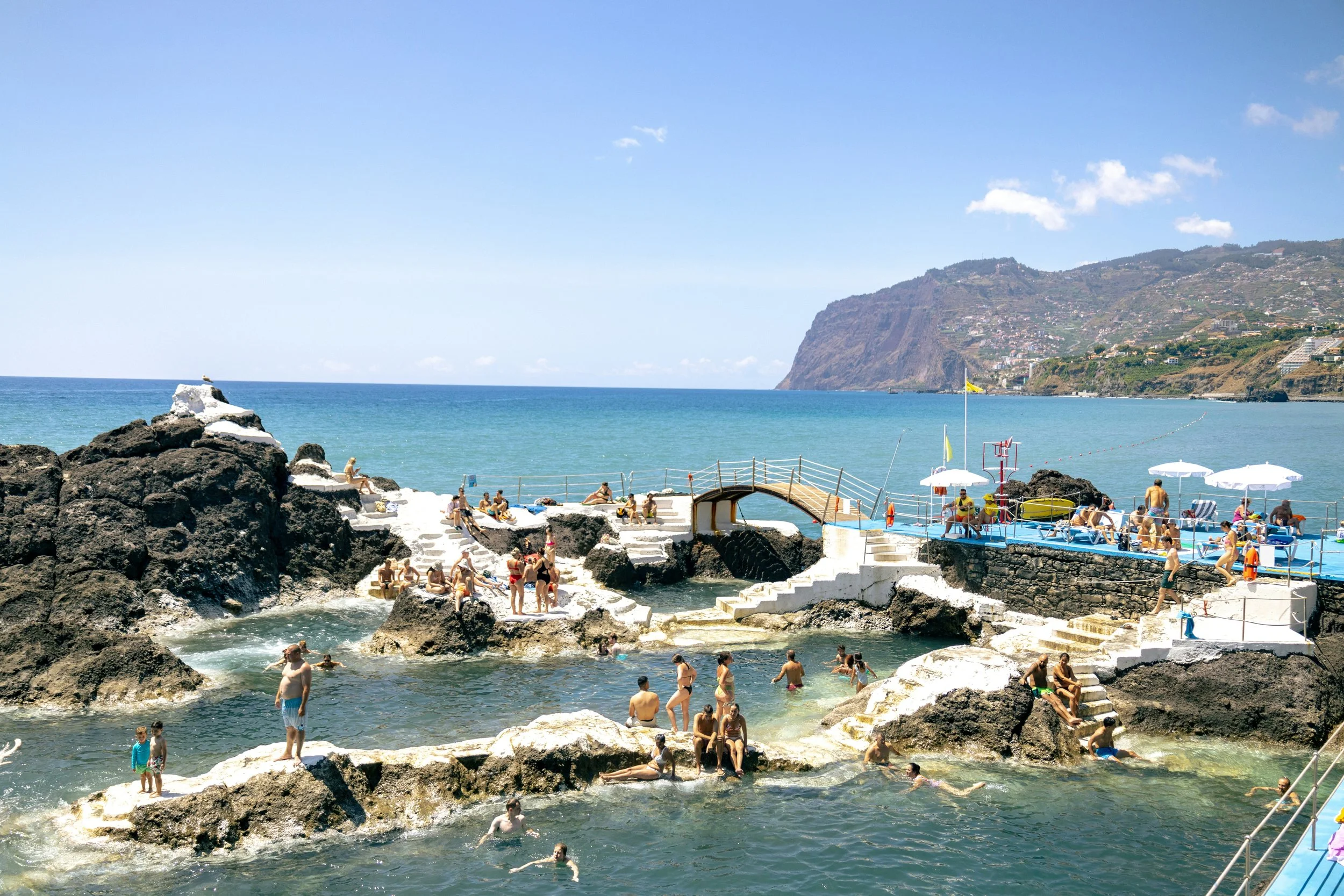 Menschen, die an einer felsigen Küste schwimmen und sich entspannen, im Hintergrund ein Strand und ein Hang unter strahlend blauem Himmel.