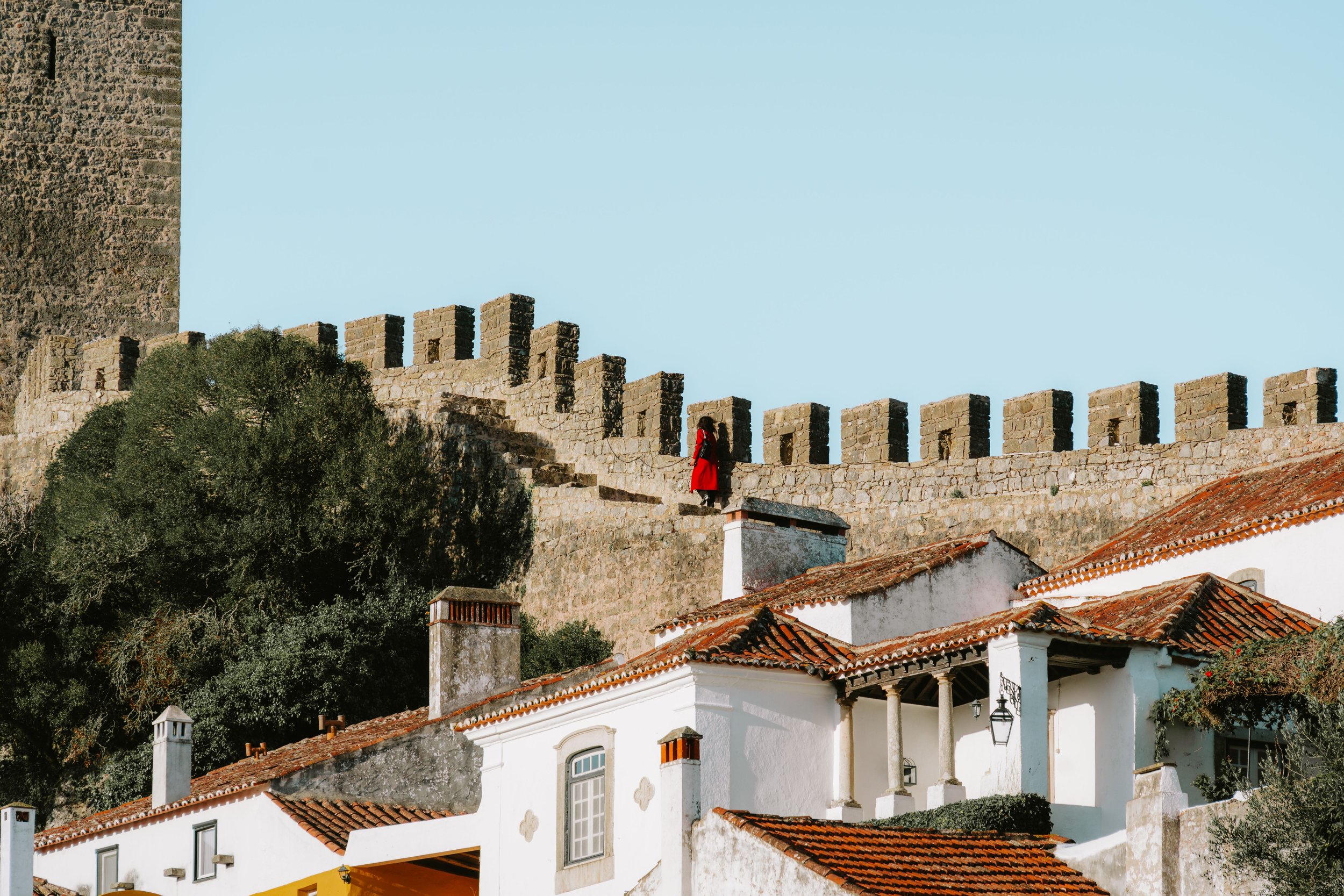 Eine Person in einem roten Mantel spaziert auf der Krone der mittelalterlichen Stadtmauer von Óbidos mit ihren Zinnen entlang, von wo aus sie auf weiße Gebäude mit Terrakottaziegeldächern blickt, umgeben von grünen Bäumen.