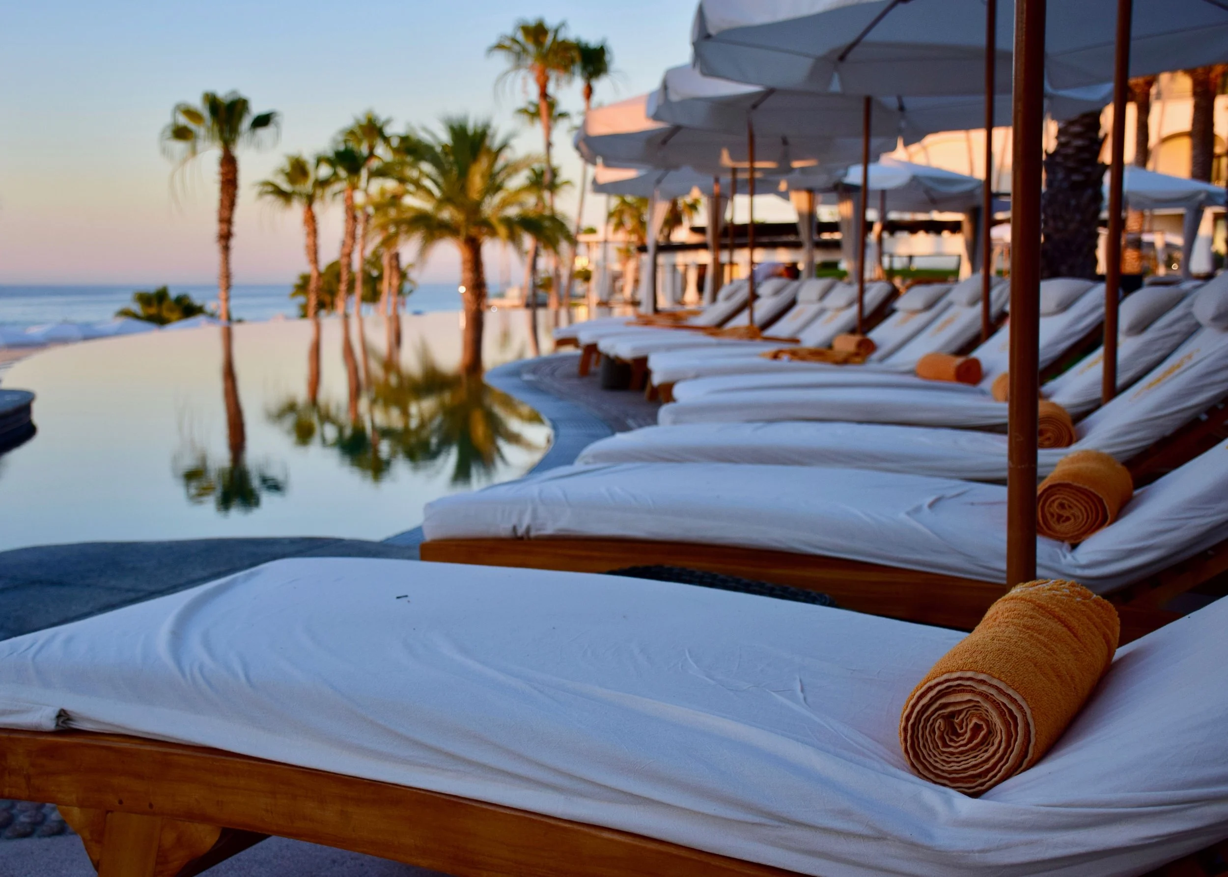 Eine Reihe weißer Liegestühle mit orangefarbenen Handtüchern und Sonnenschirmen am Poolbereich direkt am Strand, im Hintergrund Palmen und das Meer bei Sonnenuntergang.