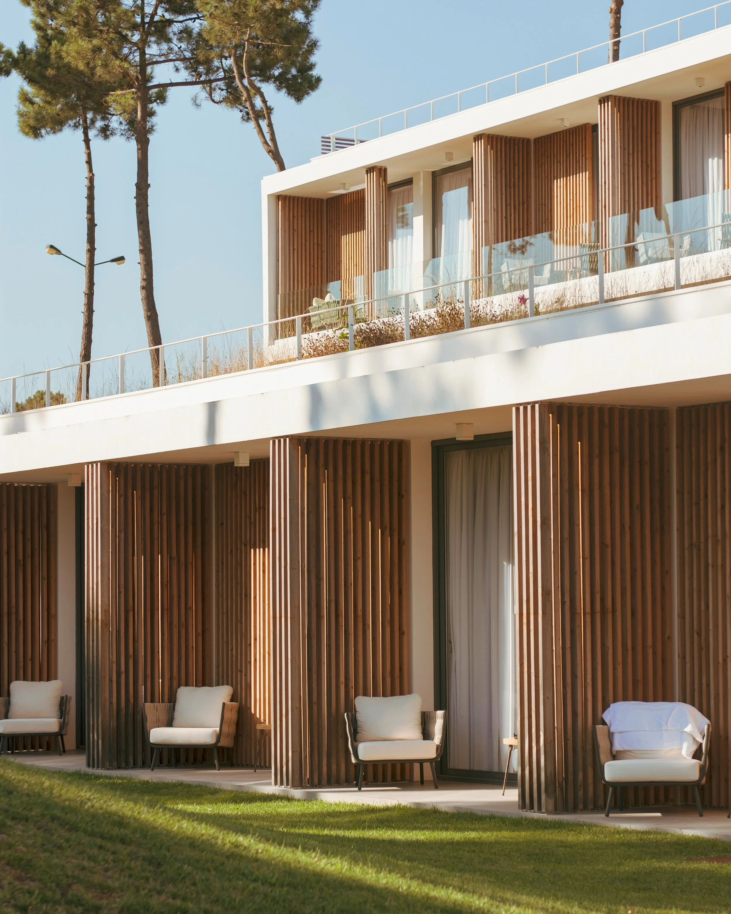 Modern residential building with large wooden slats on the exterior, glass balcony railings, and four outdoor chairs with cushions on a patio, surrounded by green grass and mature trees, under a clear blue sky.