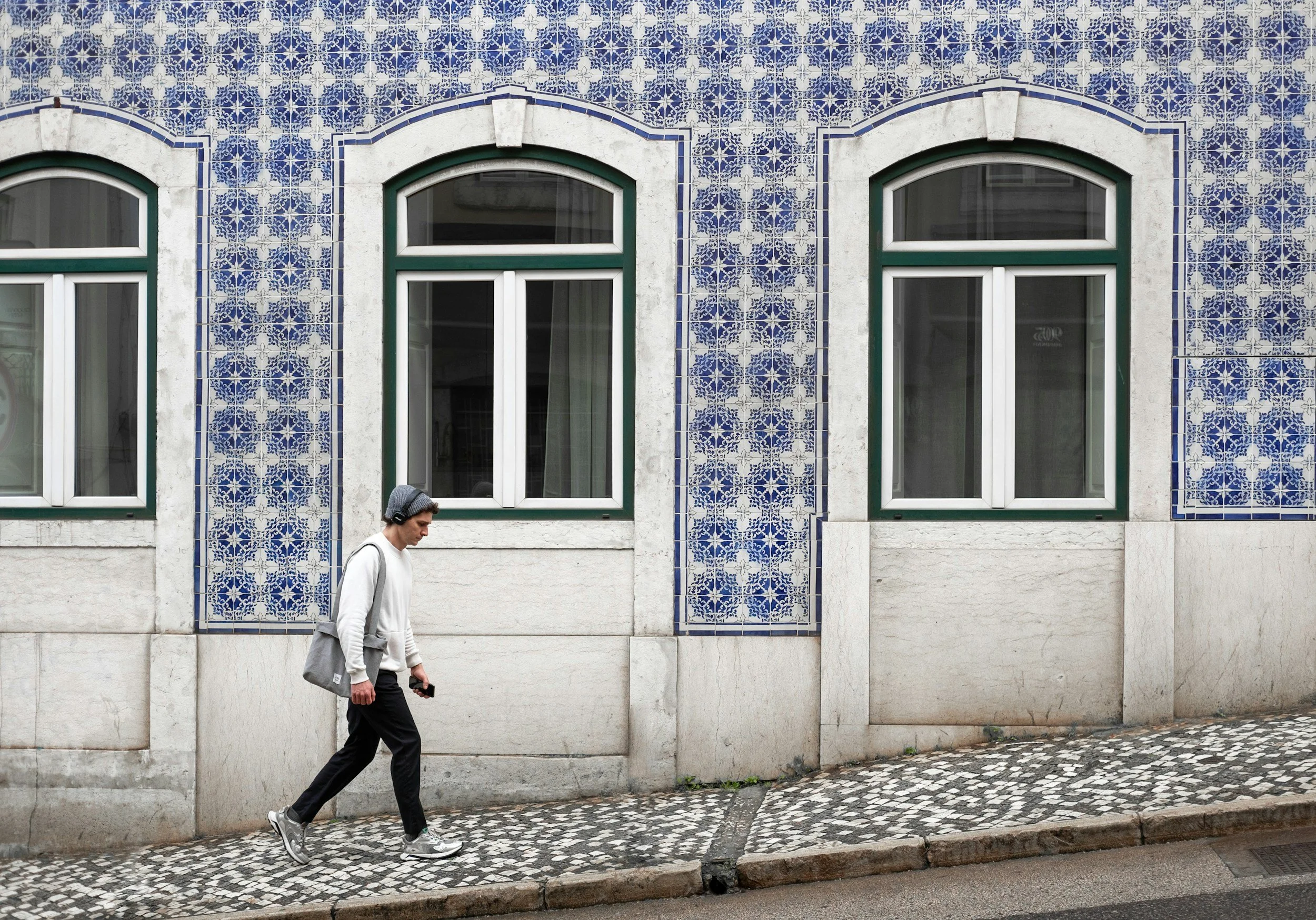 A man with gray hair, dressed in a white hoodie, black pants, gray sneakers, and carrying a gray bag, walking on a cobblestone sidewalk in front of a building with blue patterned tiles and three windows.