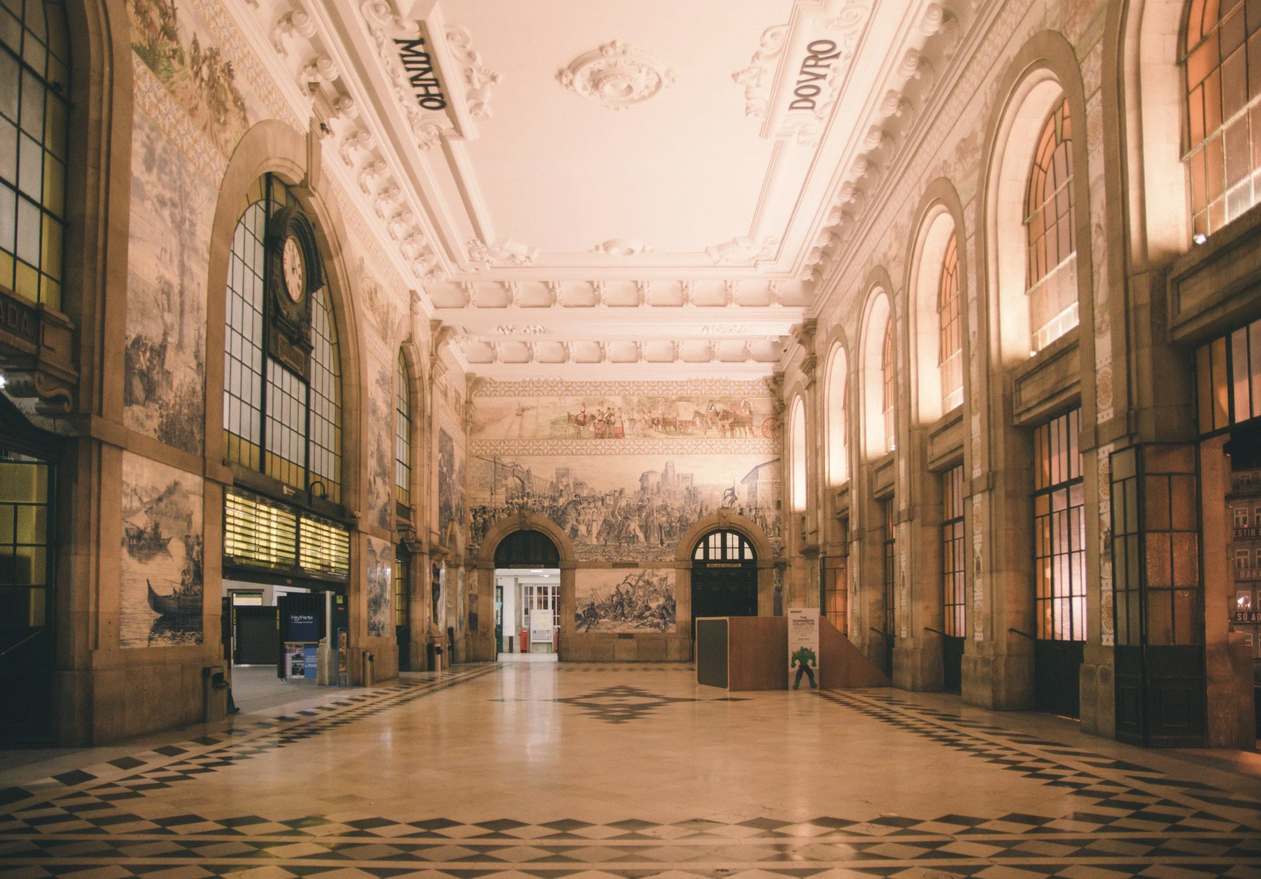 Interior of a historic train station with high arched windows, intricate decorative ceiling, mosaic tile flooring, and mural artwork on the walls.
