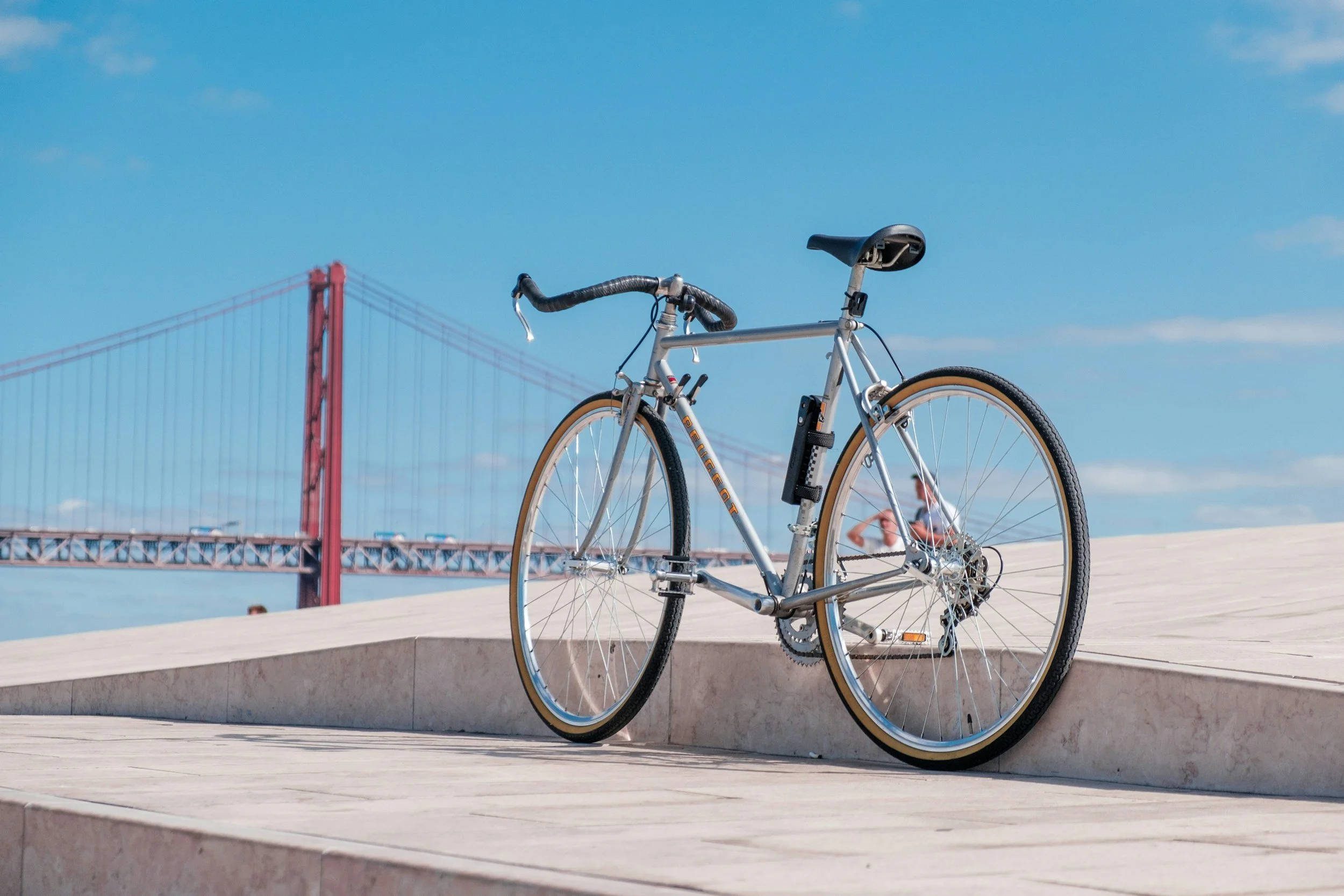 A silver bicycle parked on a concrete ledge beneath the San Francisco Golden Gate Bridge, with a bright blue sky and some clouds in the background.
