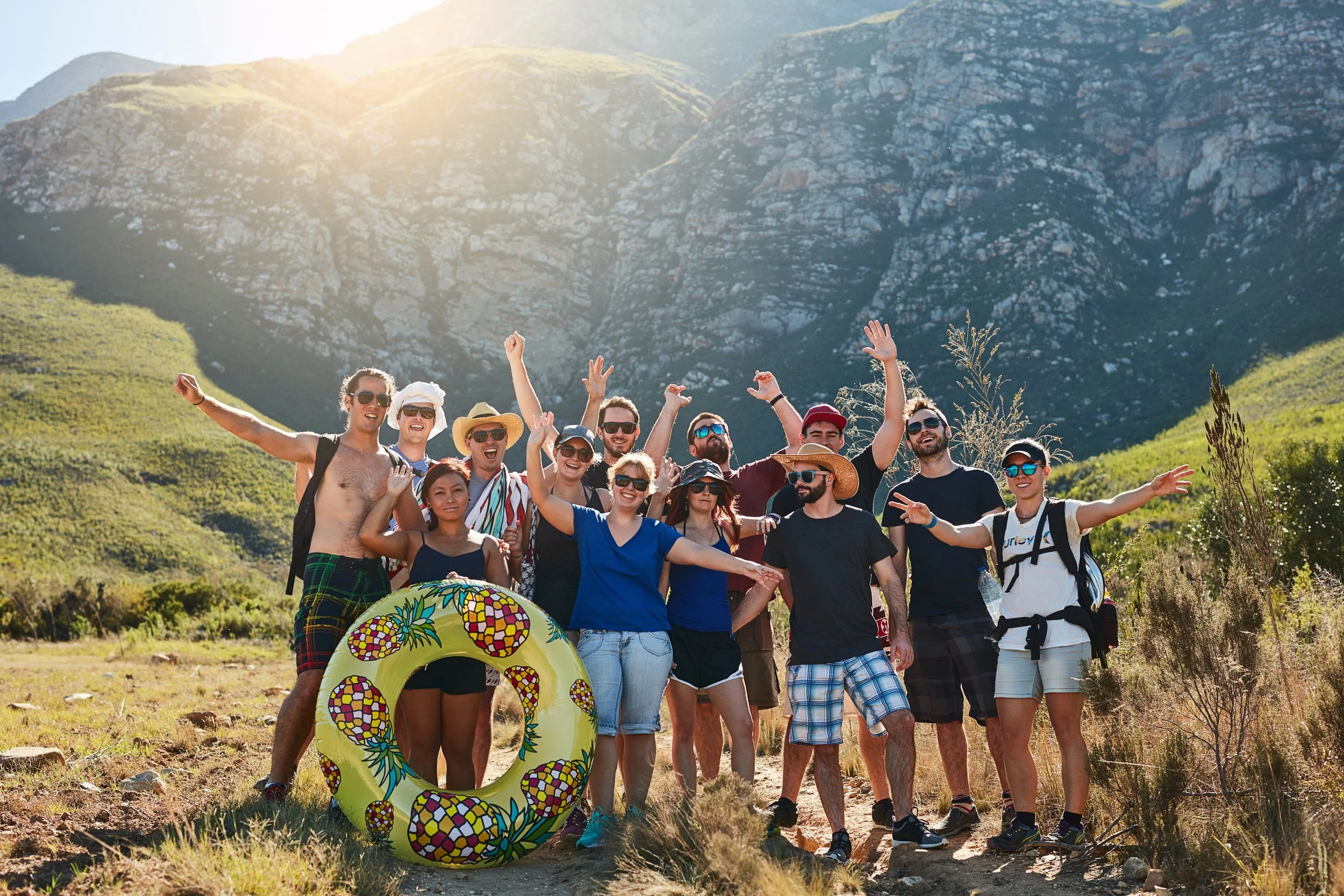 Eine Gruppe von Menschen, die an einem sonnigen Tag im Freien steht, im Hintergrund die portugiesischen Berge; einige tragen Sonnenbrillen und Hüte, lächeln und heben die Hände, im Vordergrund ist eine aufblasbare Ananas-Schwimmhilfe zu sehen.