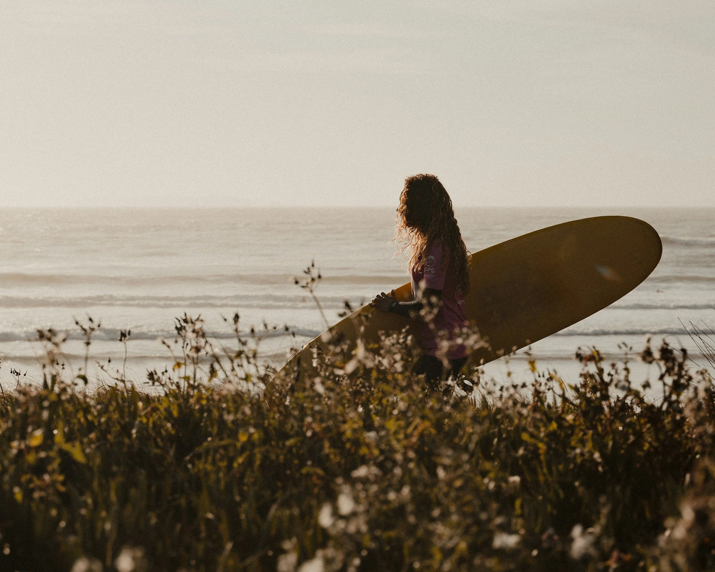 Eine Person mit lockigem Haar und einem lila Shirt, die am Strand sitzt und ein Surfbrett in der Hand hält, im Hintergrund das Meer und der Himmel.