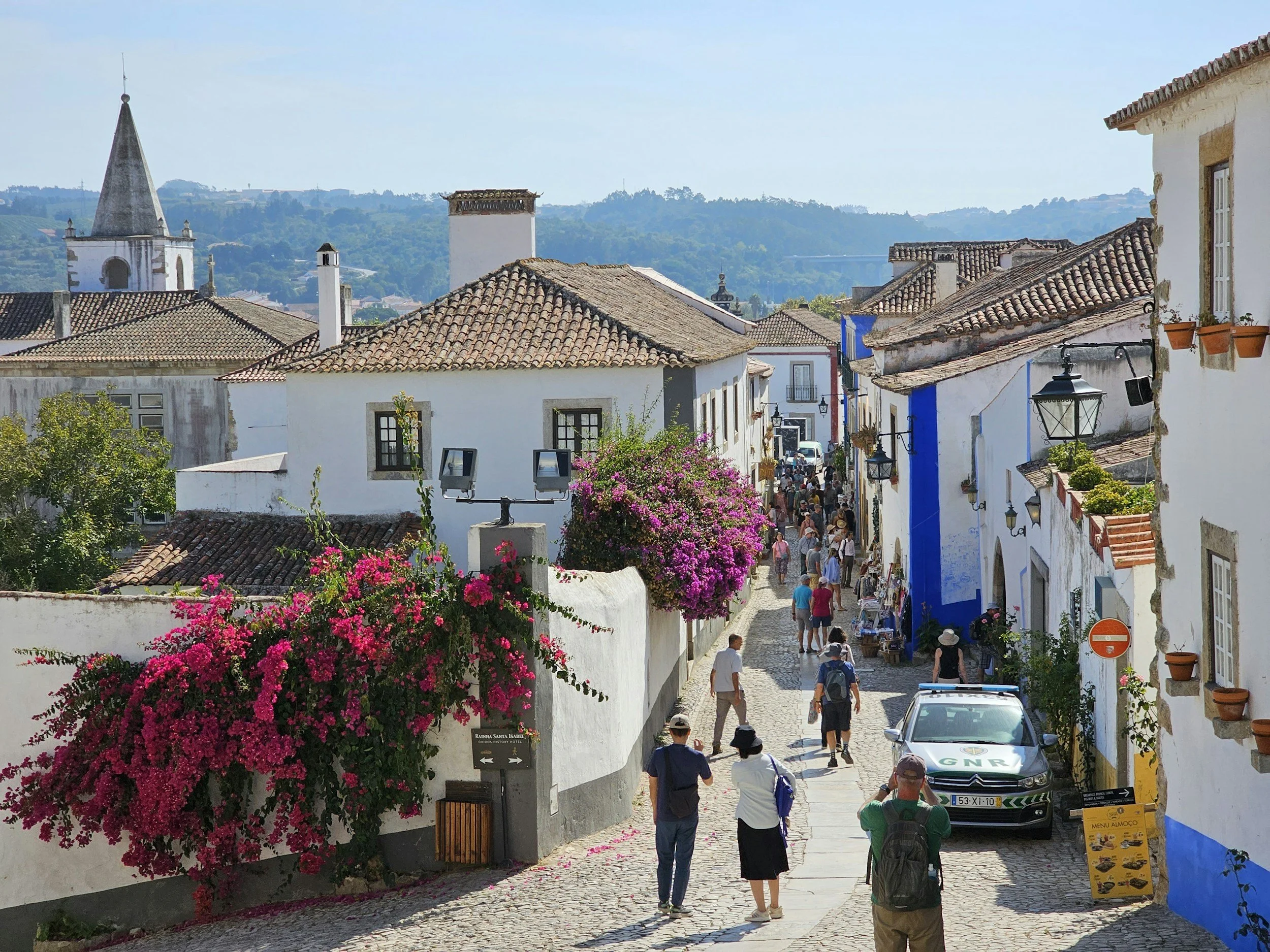 Eine belebte Straße in einer Stadt am Mittelmeer mit weißen Gebäuden, Terrakottadächern und leuchtend rosa Bougainvillea-Blüten. Die Menschen spazieren und gehen einkaufen, im Hintergrund ist vor einer üppigen Hügellandschaft ein Kirchturm zu sehen.