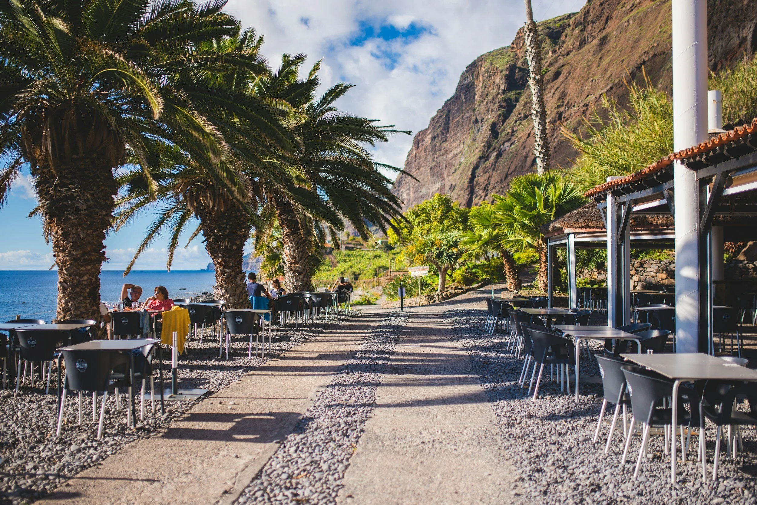 Ein Restaurant im Freien am Meer mit Tischen und Stühlen, hohen Palmen, einem Berg im Hintergrund und einem teilweise bewölkten Himmel.