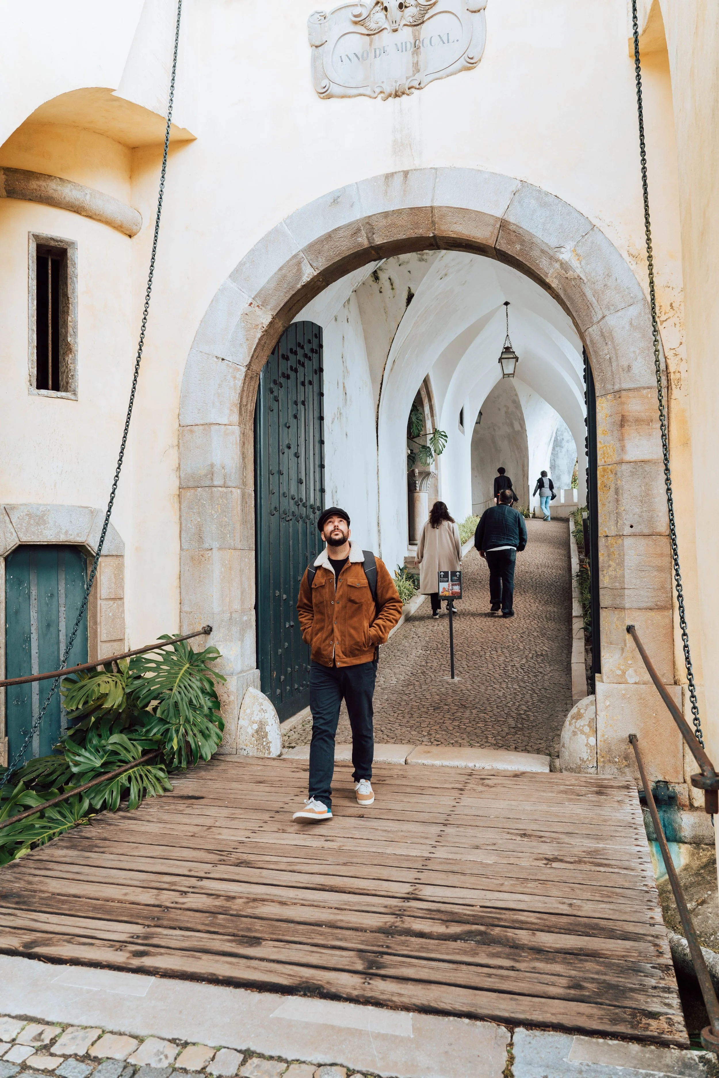 A man with a beard and hat walking on a wooden bridge through an arched stone entranceway, with other people walking in the background and greenery on the sides.