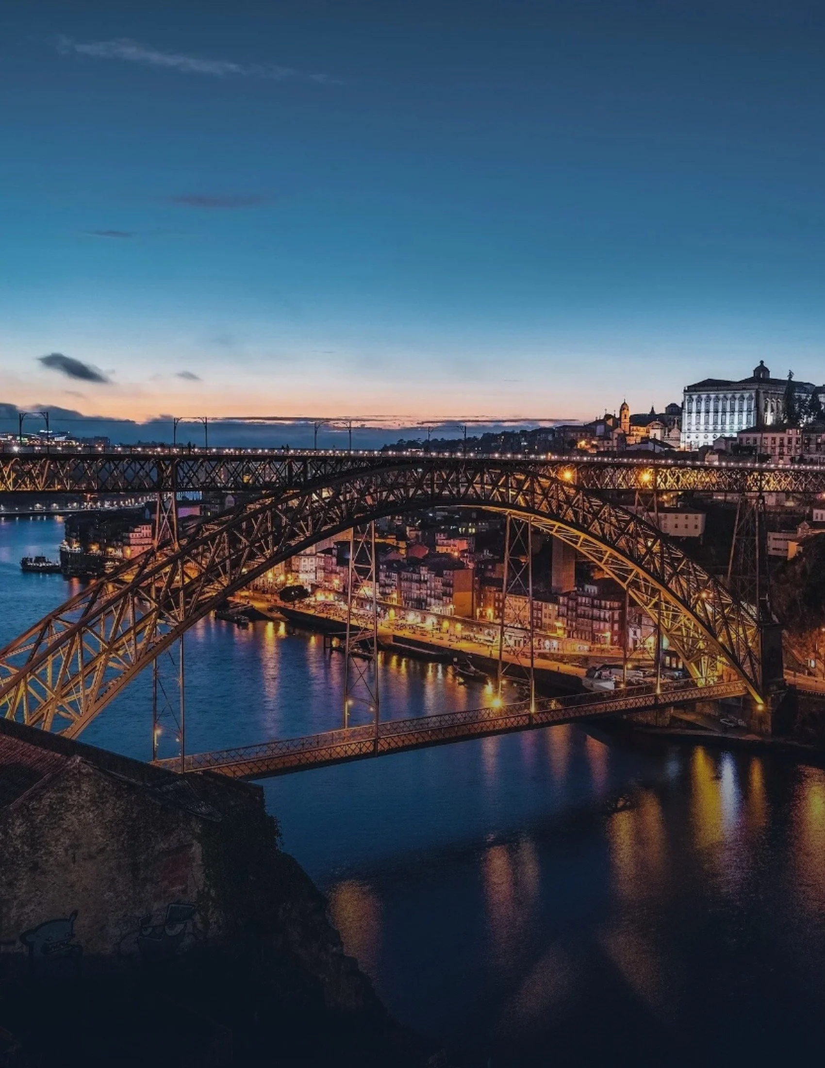 A cityscape at dusk featuring a bridge over a river, with buildings and a church or cathedral with a tall spire in the background, and the sky transitioning from sunset colors to night.
