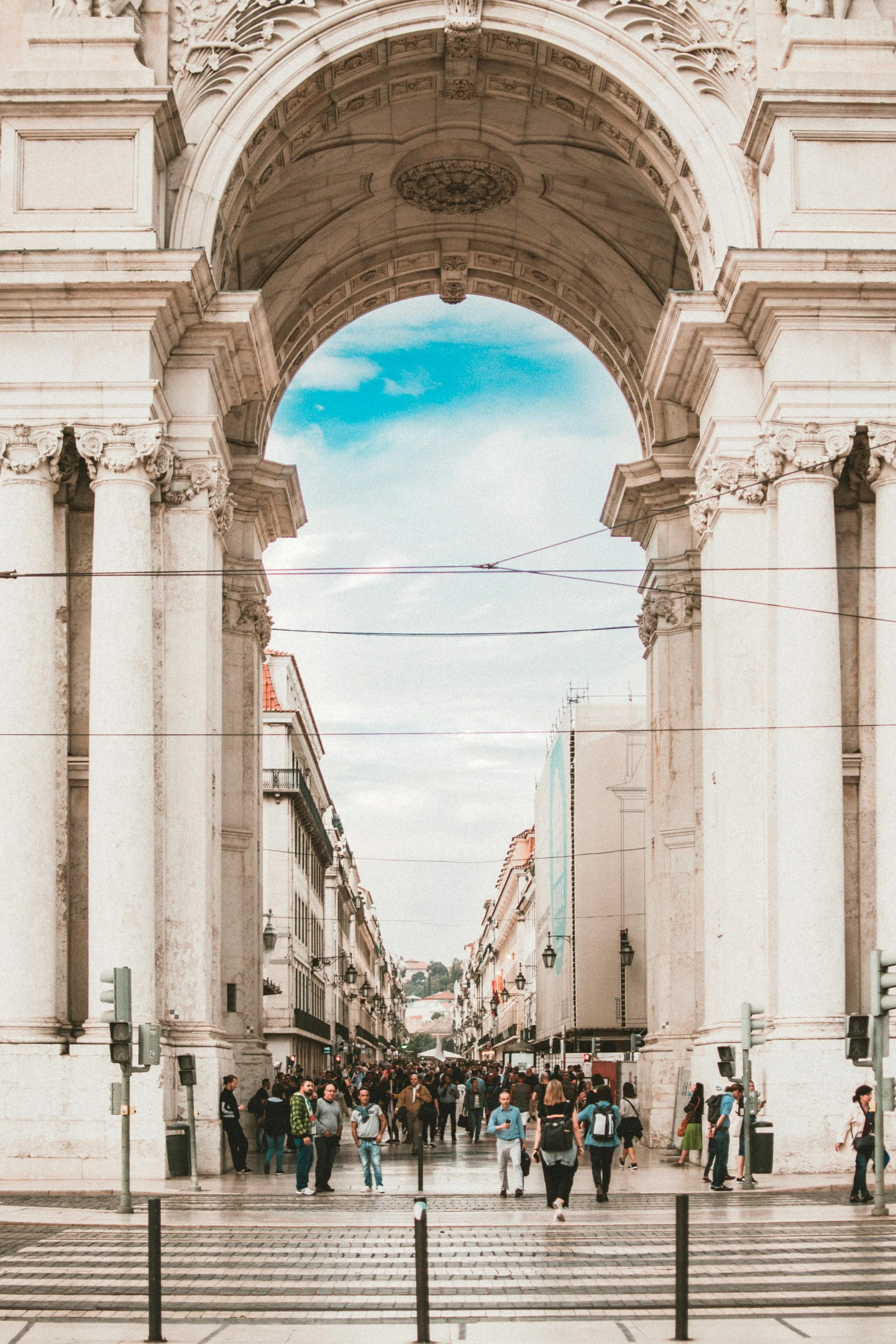 A busy city street viewed through a large, ornate archway with white columns and detailed carvings. Pedestrians are walking and gathering on the street, with buildings and streetlights lining the sides. The sky is partly cloudy.