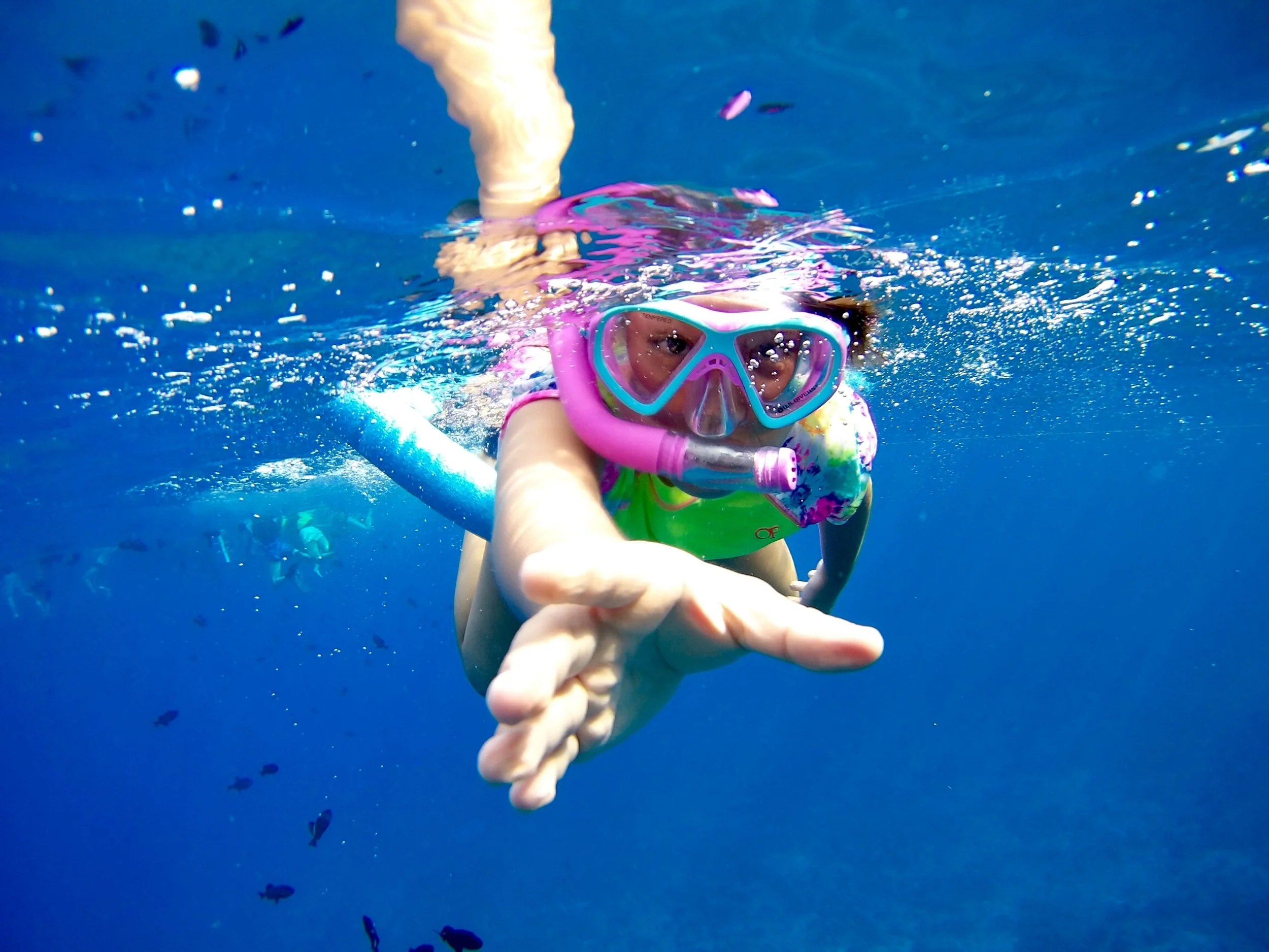 Ein junges Mädchen mit Schnorchelmaske und Schwimmshirt, das unter Wasser schwimmt und die Hand ausstreckt.
