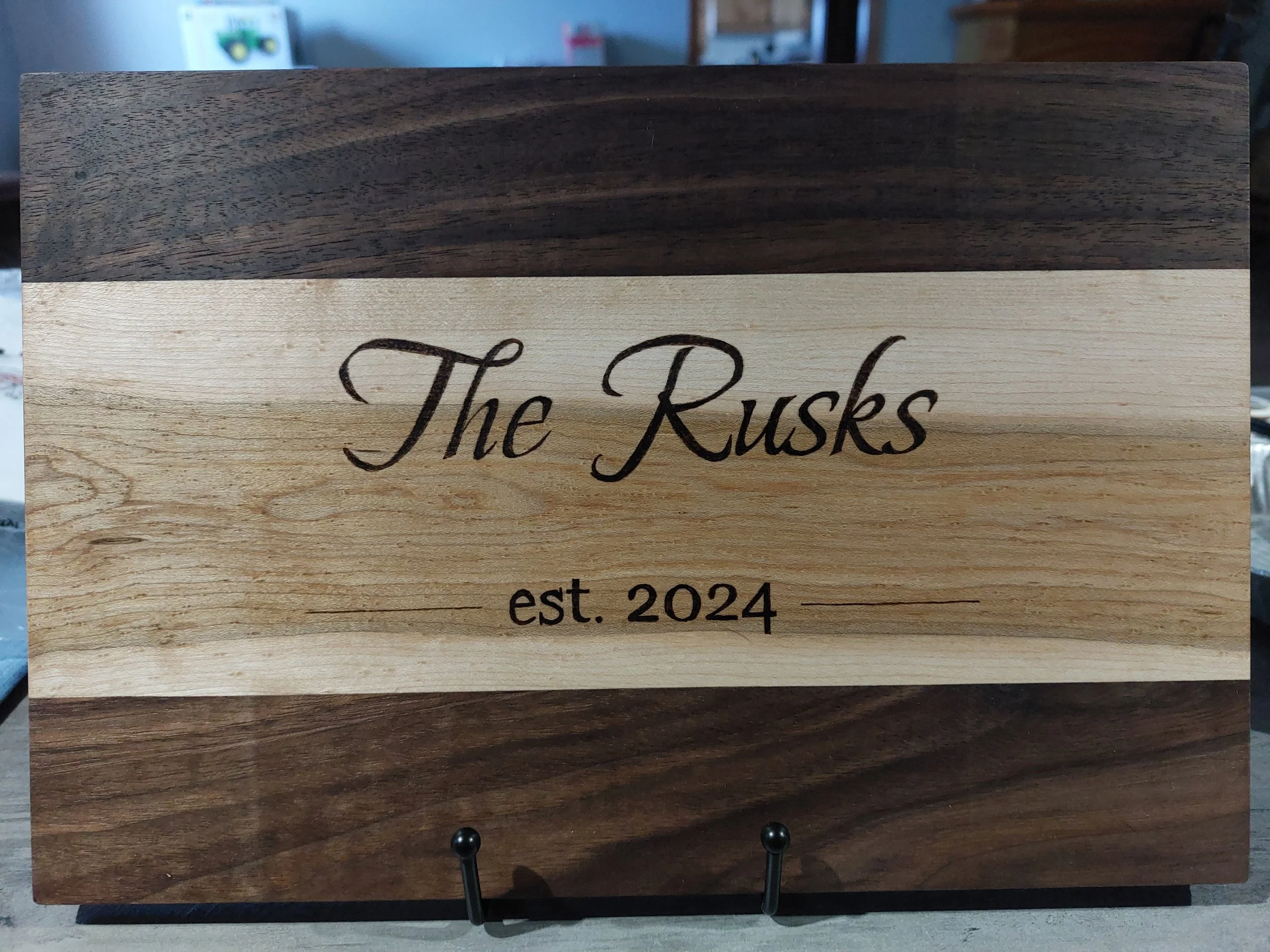 A wooden cutting board with a light-colored central strip and dark-colored top and bottom sections. The plaque has the words "The Rusks" burned in cursive and "est. 2024" in smaller print below, with two horizontal lines on either side of the year.