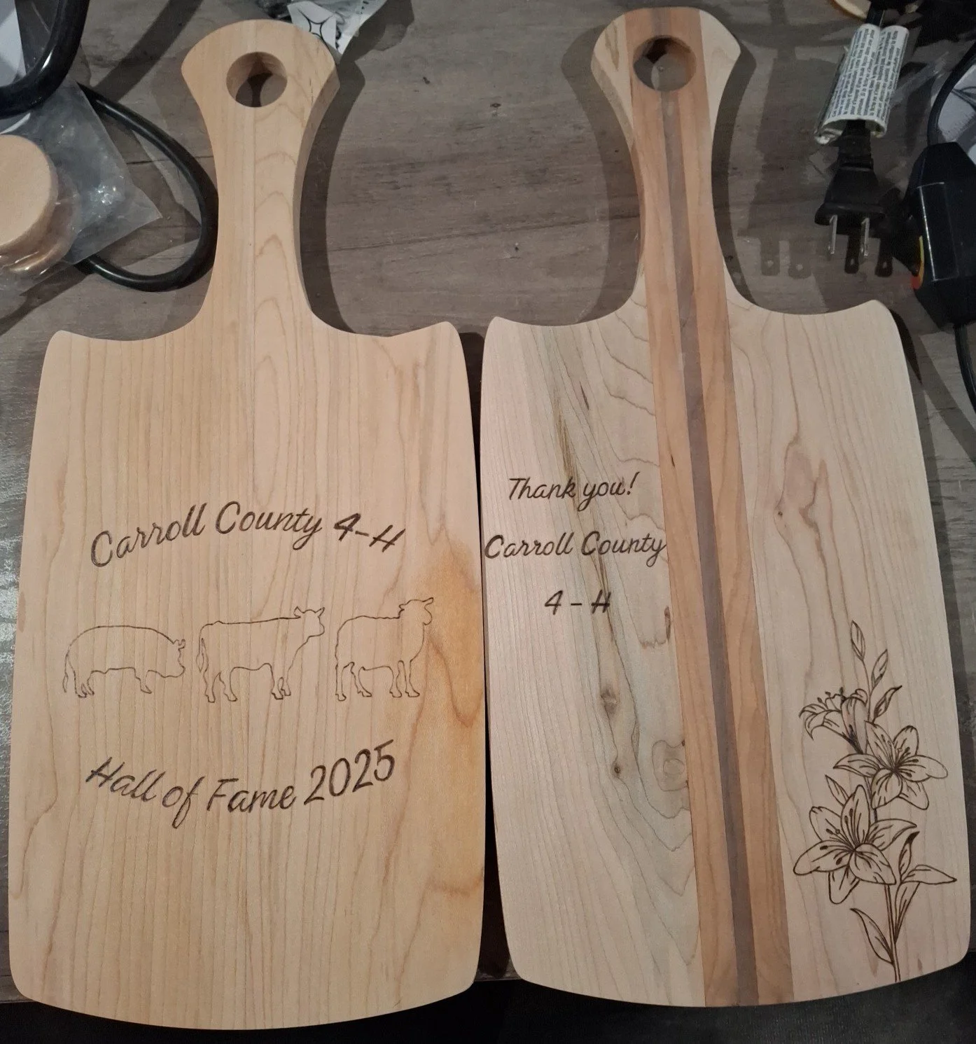 Two wooden cutting boards, one with burned cows, text, and floral design, and one with burned with text and flowers, placed on a table with miscellaneous items around.