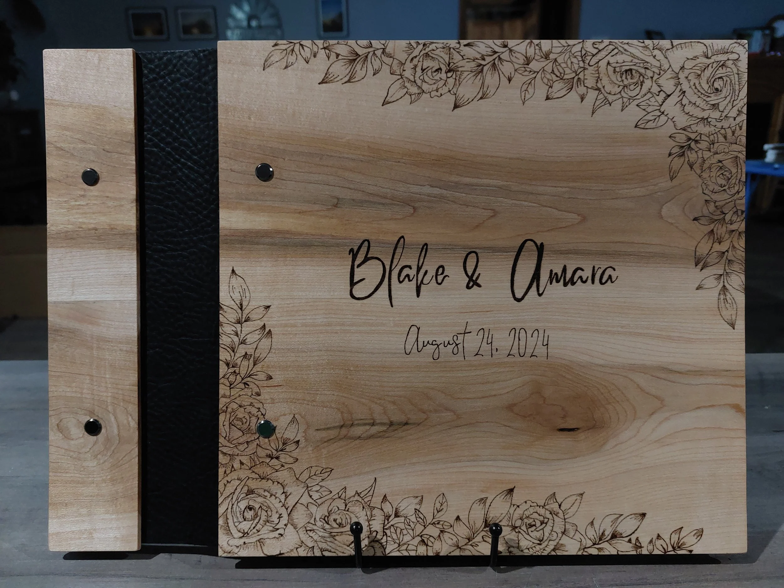 Wooden wedding guest book with hand burned rose and leaf border, displaying the names Blake and Amara and the date August 24, 2024.