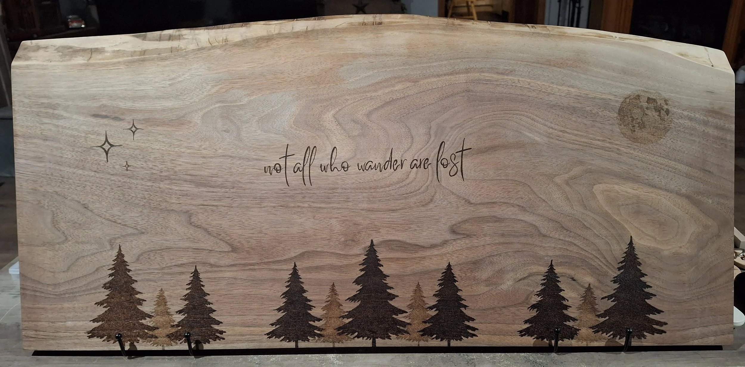 Decorative wooden charcuterie board featuring the quote 'not all who wonder are lost' written in cursive, with illustrations of trees at the bottom, stars and a moon in the sky.