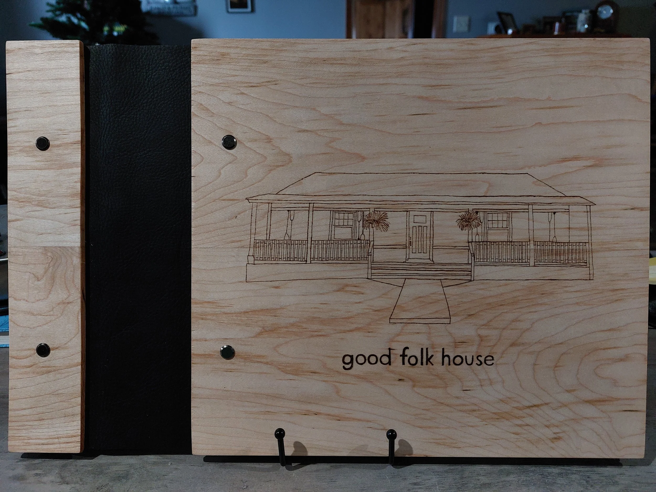 A wooden guest book with a line burning of a house showing front porch, stairs, and two potted plants, with the caption 'good folk house' at the bottom.