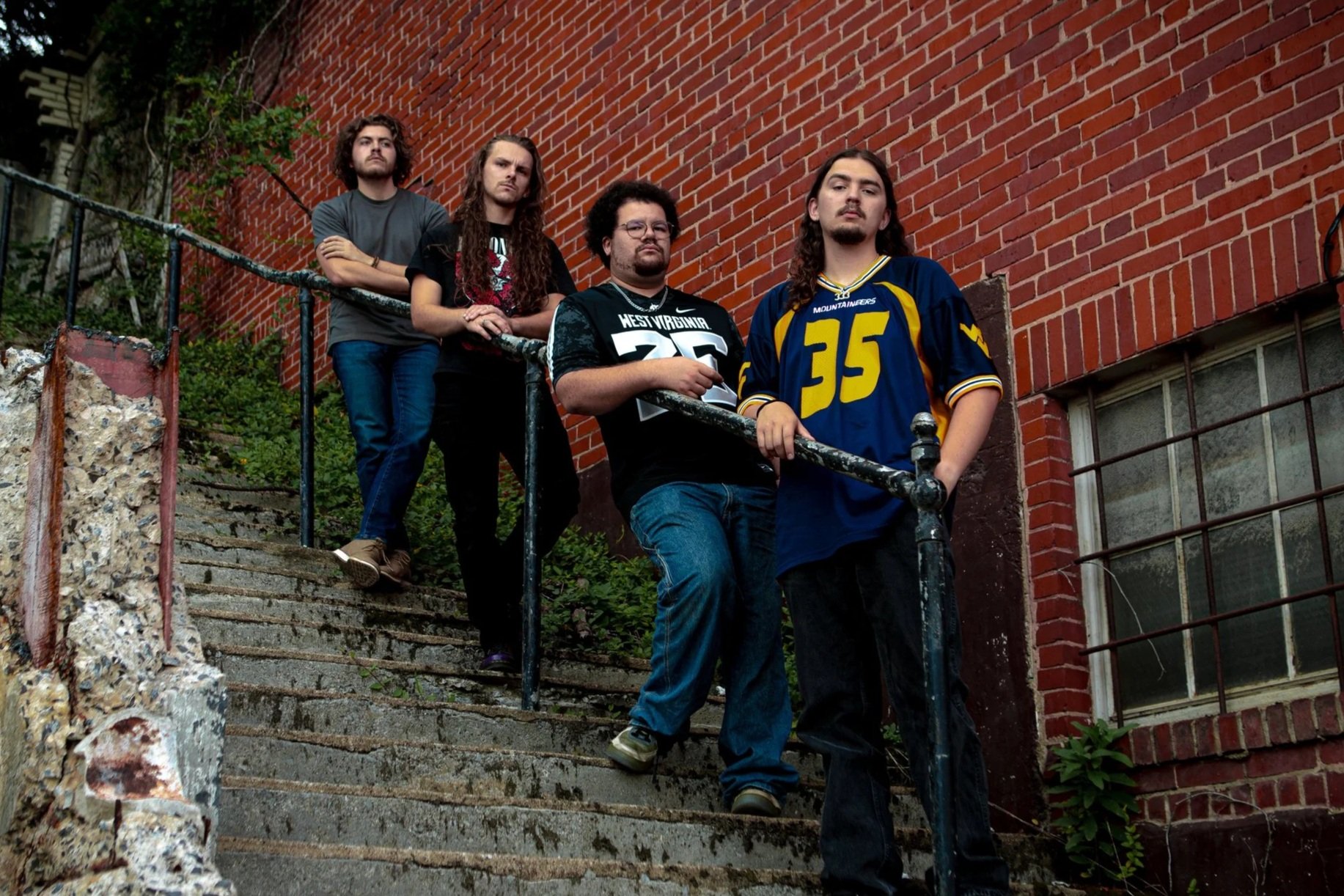 Five young men standing on outdoor concrete stairs beside a brick building. Some are wearing sports jerseys, with a railing and greenery in the background.