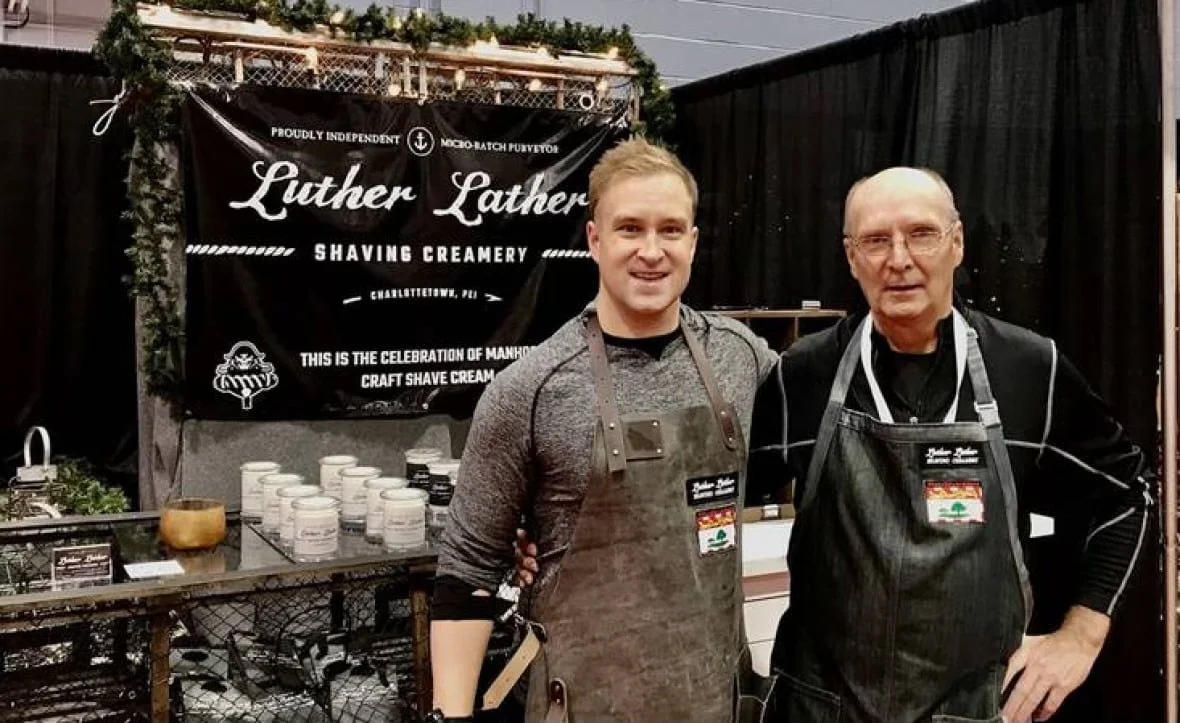 Two men wearing aprons standing together in front of a black sign at a shaved creamery booth. The sign reads 'Luther Lather Shaving Creamery.'
