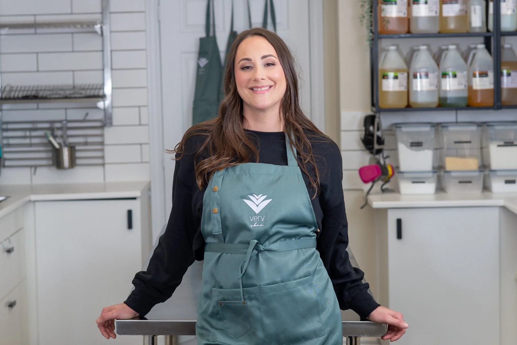 A woman with long brown hair wearing an apron and black shirt, standing in an indoor space with shelves filled with jars and containers.