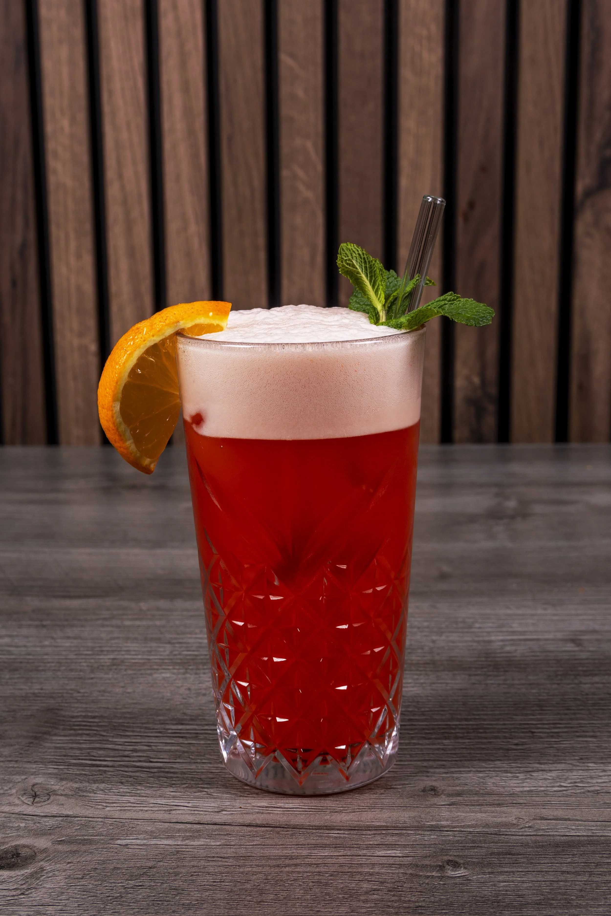 Een glas met een roze cocktail, geserveerd met een schijfje sinaasappel en muntblaadjes, op een houten tafel met een houten achtergrond.