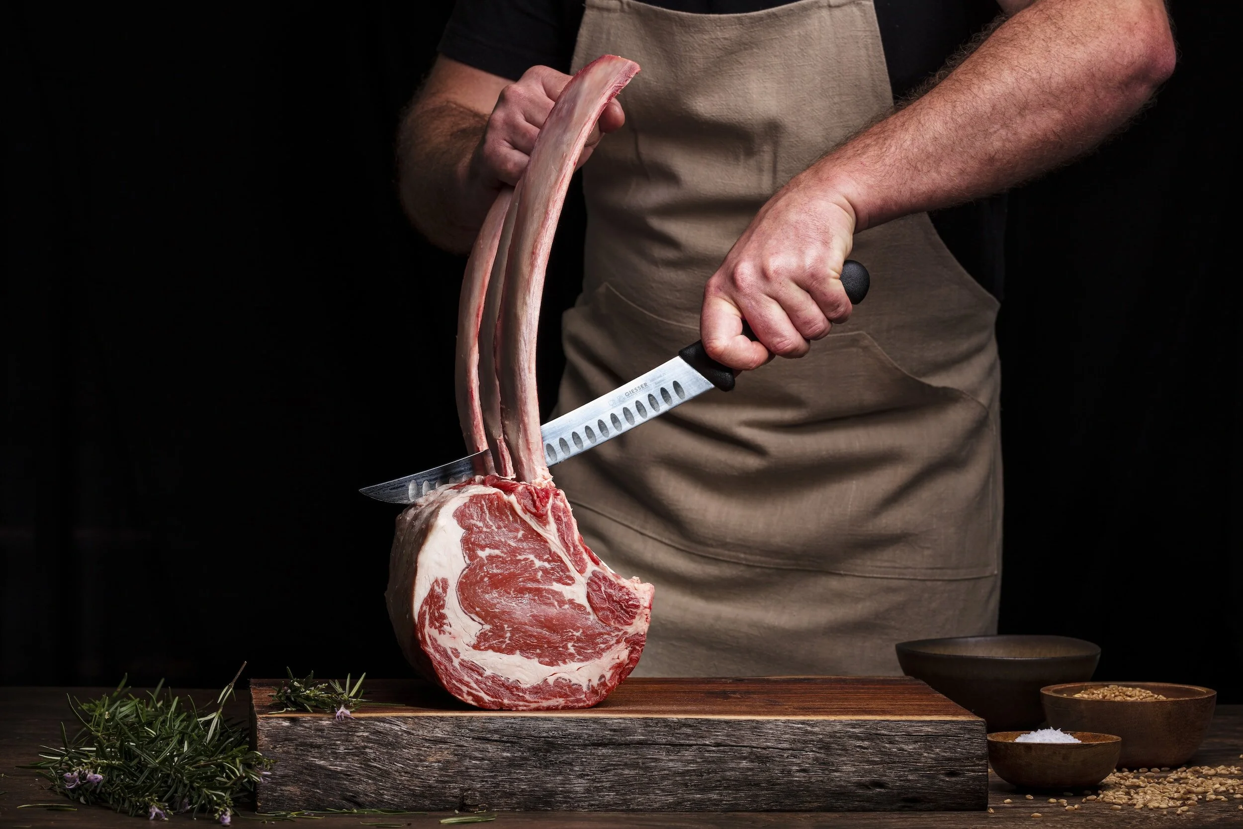 Beenhouwer snijdt een stuk rundvlees (côte á l'os) met drie ribben met een groot mes, op een houten snijplank, met kruiden en kommen op de tafel.