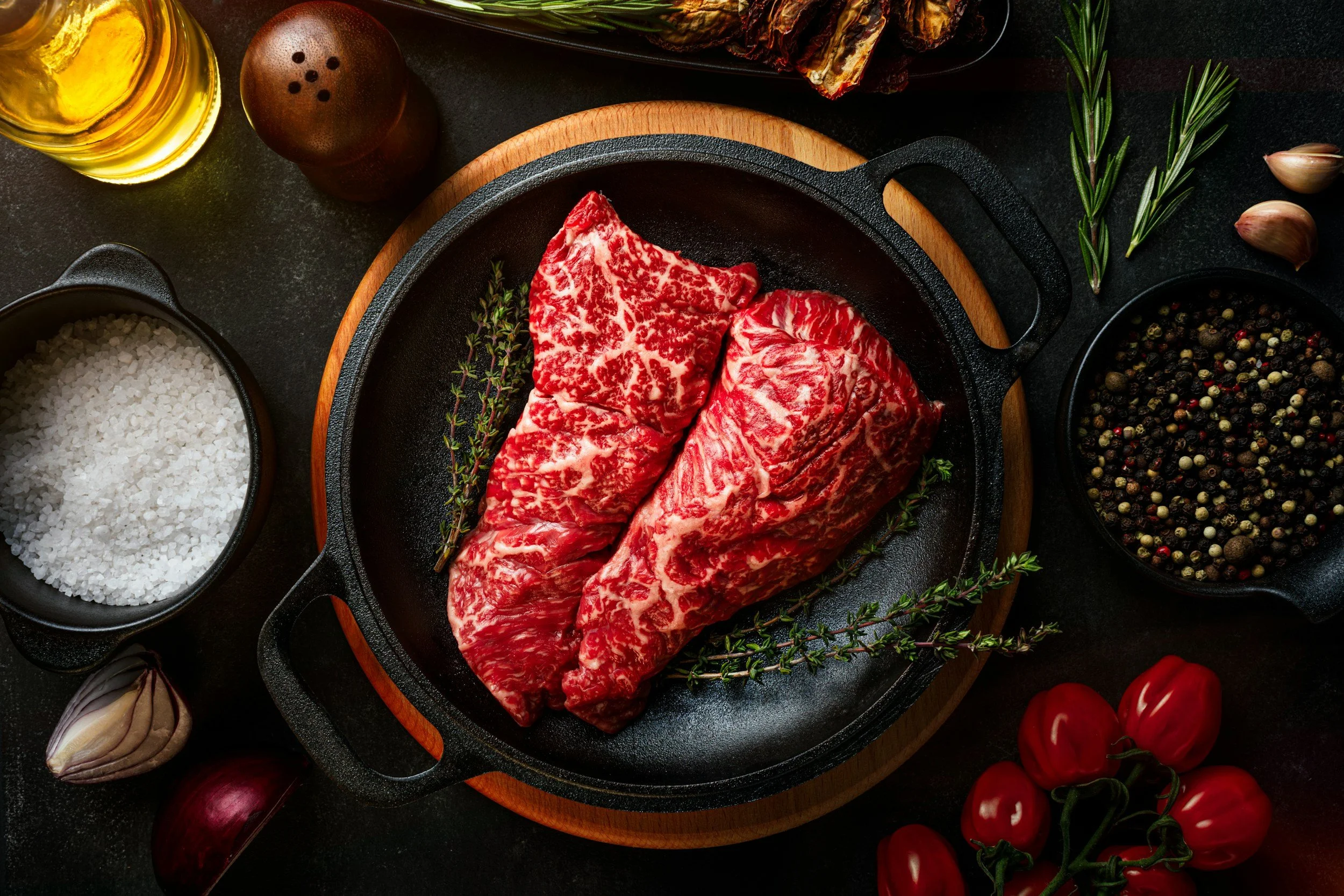 Verschillende ingrediënten voor het bereiden van vlees, inclusief gemarmerde steaks in een pan, met kruiden, olijfolie, zout, peper en groenten rondom, op een donkere achtergrond.