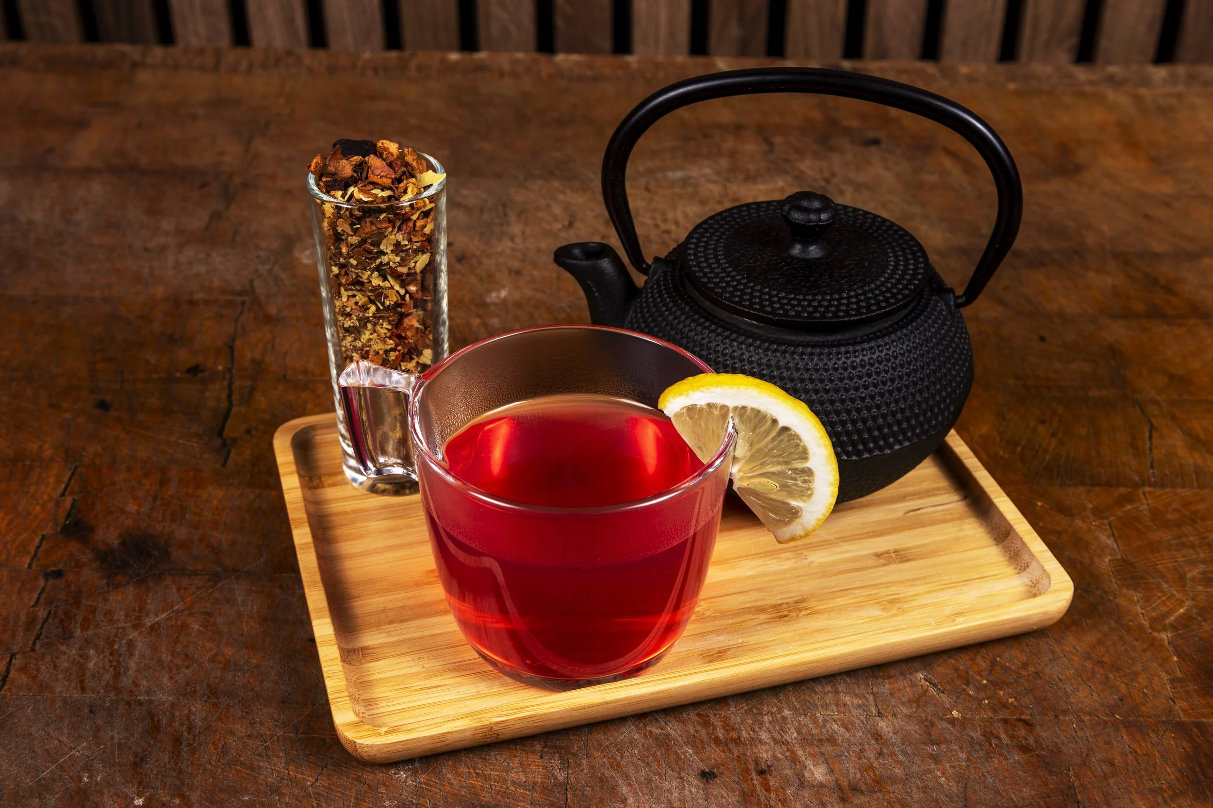 Een glas met roodgekleurde thee, een schijfje citroen, een zwarte theepot, en een glas met losse thee kruiden op een houten dienblad op een houten tafel.