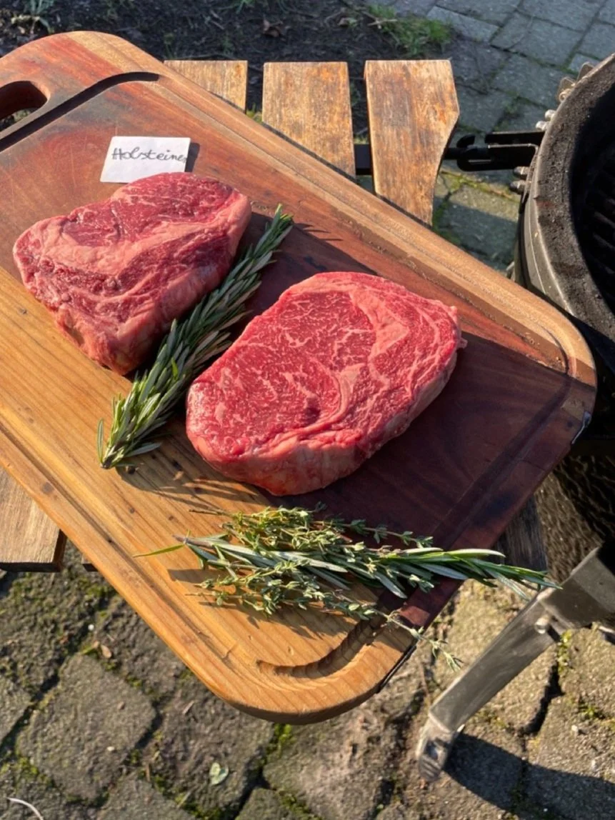 Twee stukken rauw vlees op een houten snijplank, versierd met een takje rozemarijn en tijm, buiten naast een BBQ.