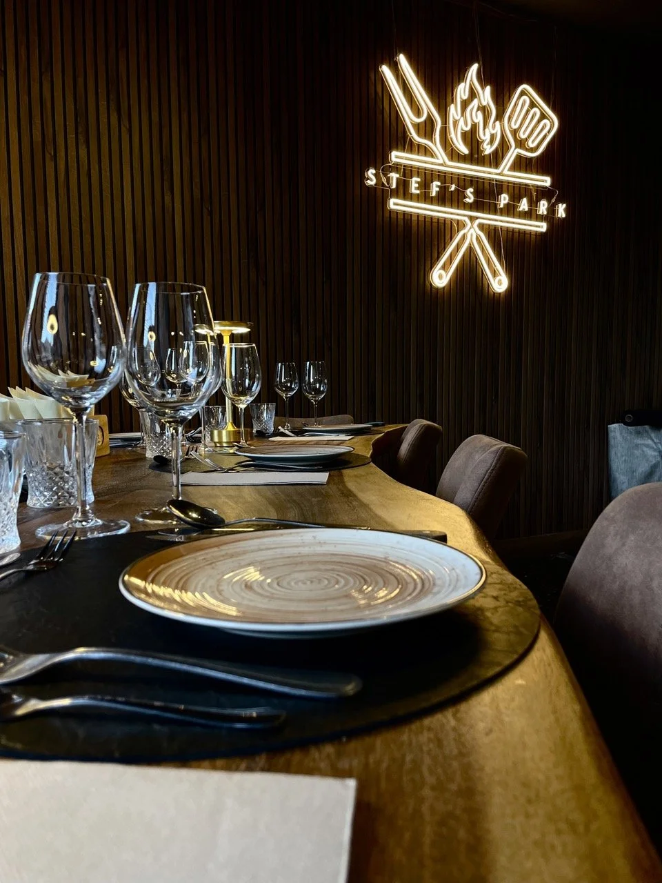Elegante restauranttafel met tomeloos glas, borden, bestek en servetten, met een houten muur en neonreclame met grill-, vork- en brandersymbool op de achtergrond.