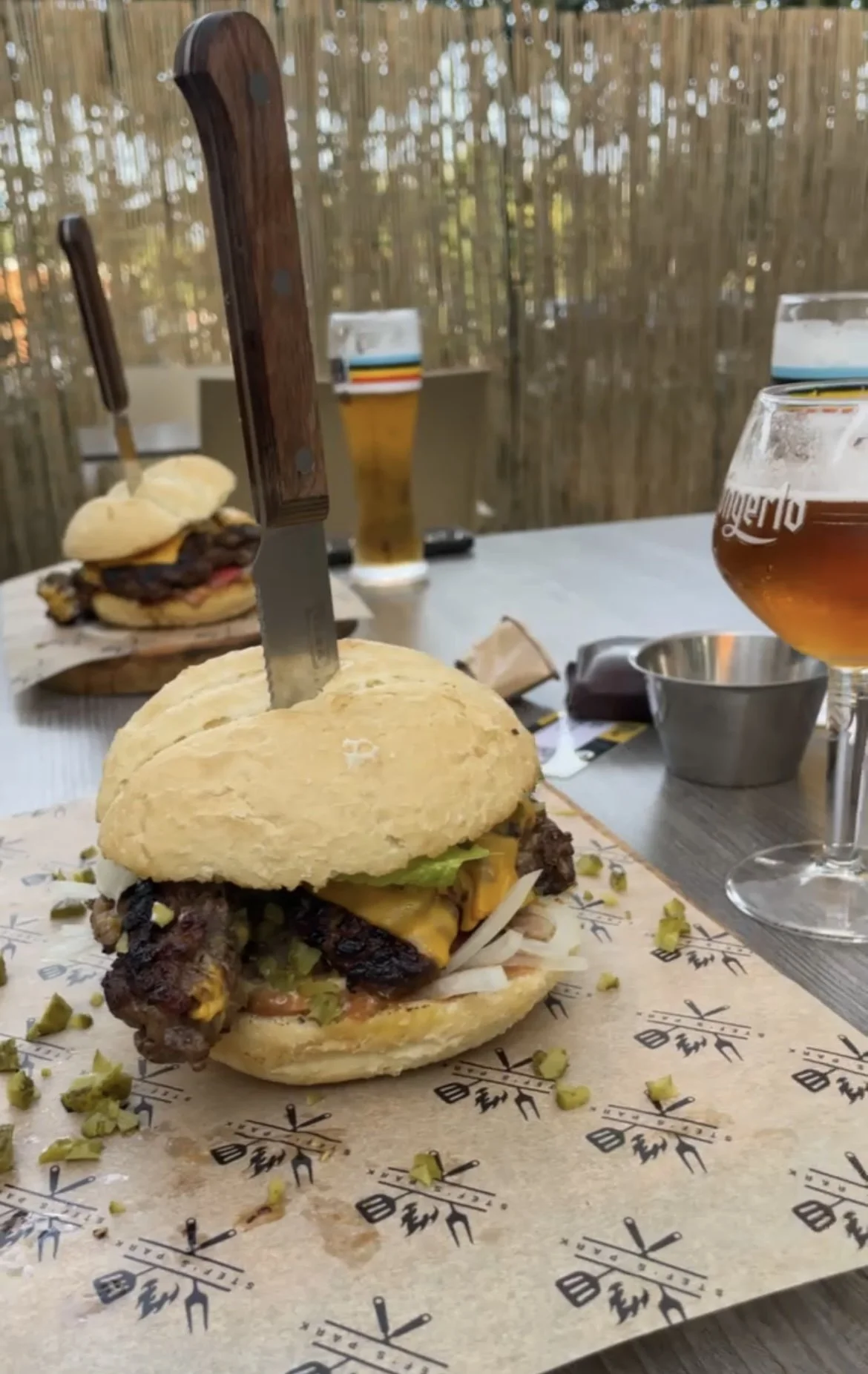 Close-up van een bewerkzame hamburger met een scherp mes erin, op een plaatje met een grafisch patroon, met een ander hamburger, bier en glazen op de achtergrond op een houten tafel buiten op het terras.