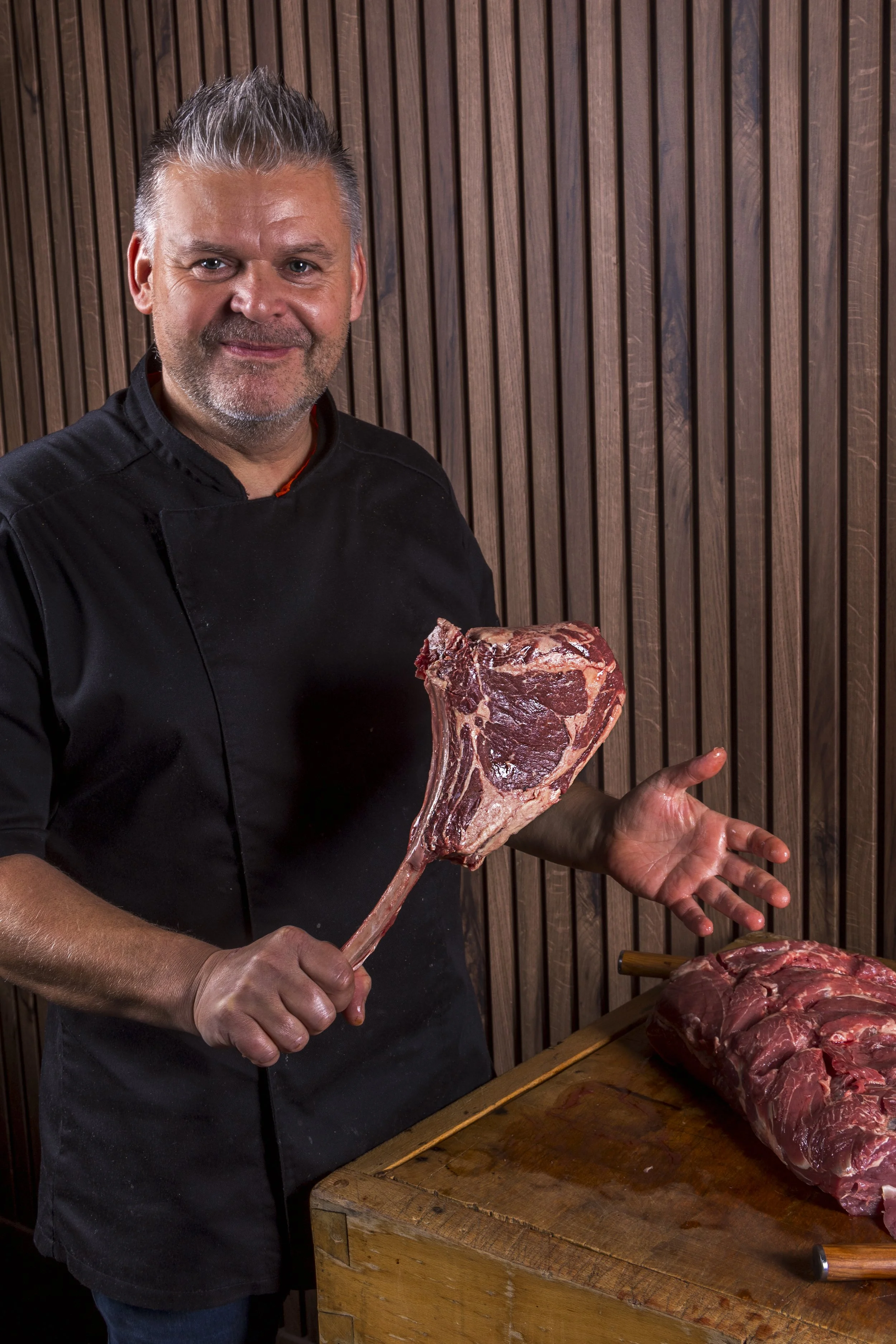 Man met een zwart koksvest met vlees op een houten tafel, houdt een grote côte á l'os vast en staat voor een houten achtergrond.