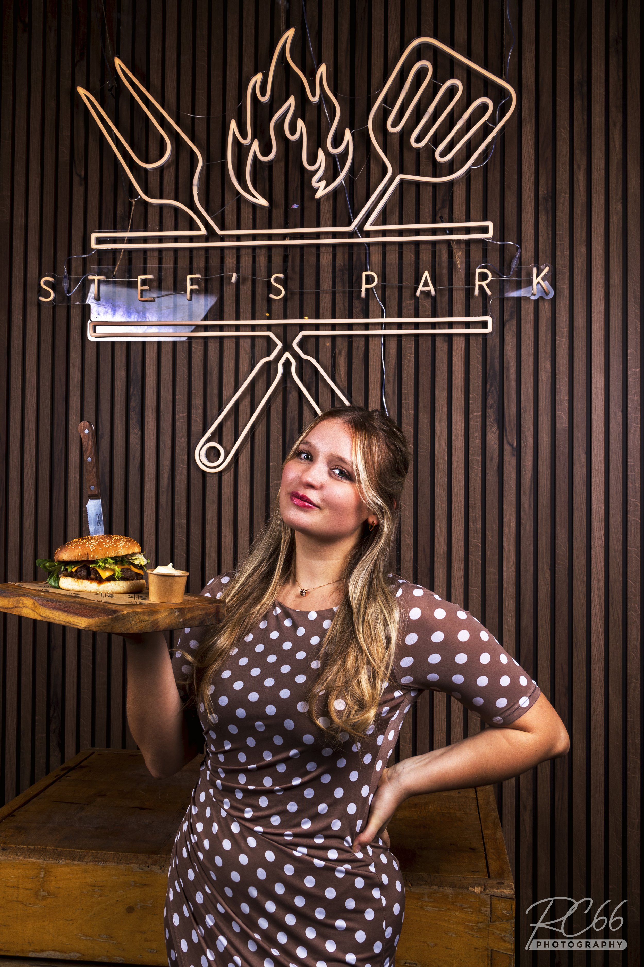 Vrouw in een polkadot jurk houdt een houten dienblad met een hamburger vast. Ze staat voor een houten achtergrond.