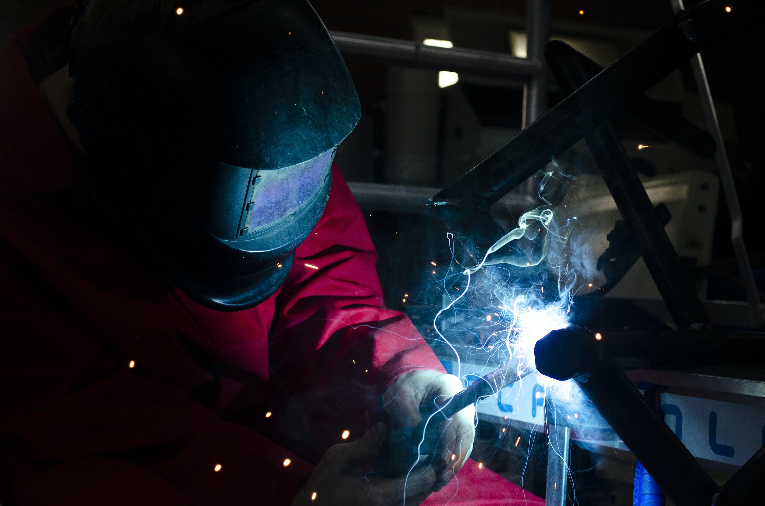 A person welding metal, wearing a protective helmet with a reflective visor and a red protective jacket, sparks and bright blue welding light surround them in a dark workshop.
