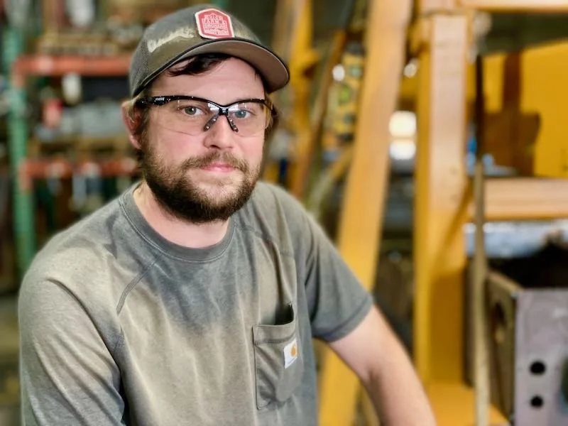 A man with glasses and a beard wearing a Carhartt t-shirt and a cap, standing in a warehouse or industrial setting with yellow metal support beams and shelves in the background.