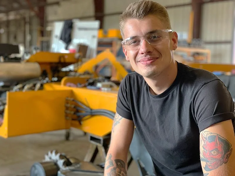 Young man with tattoos and glasses smiling in a workshop with yellow industrial equipment and tools in the background.