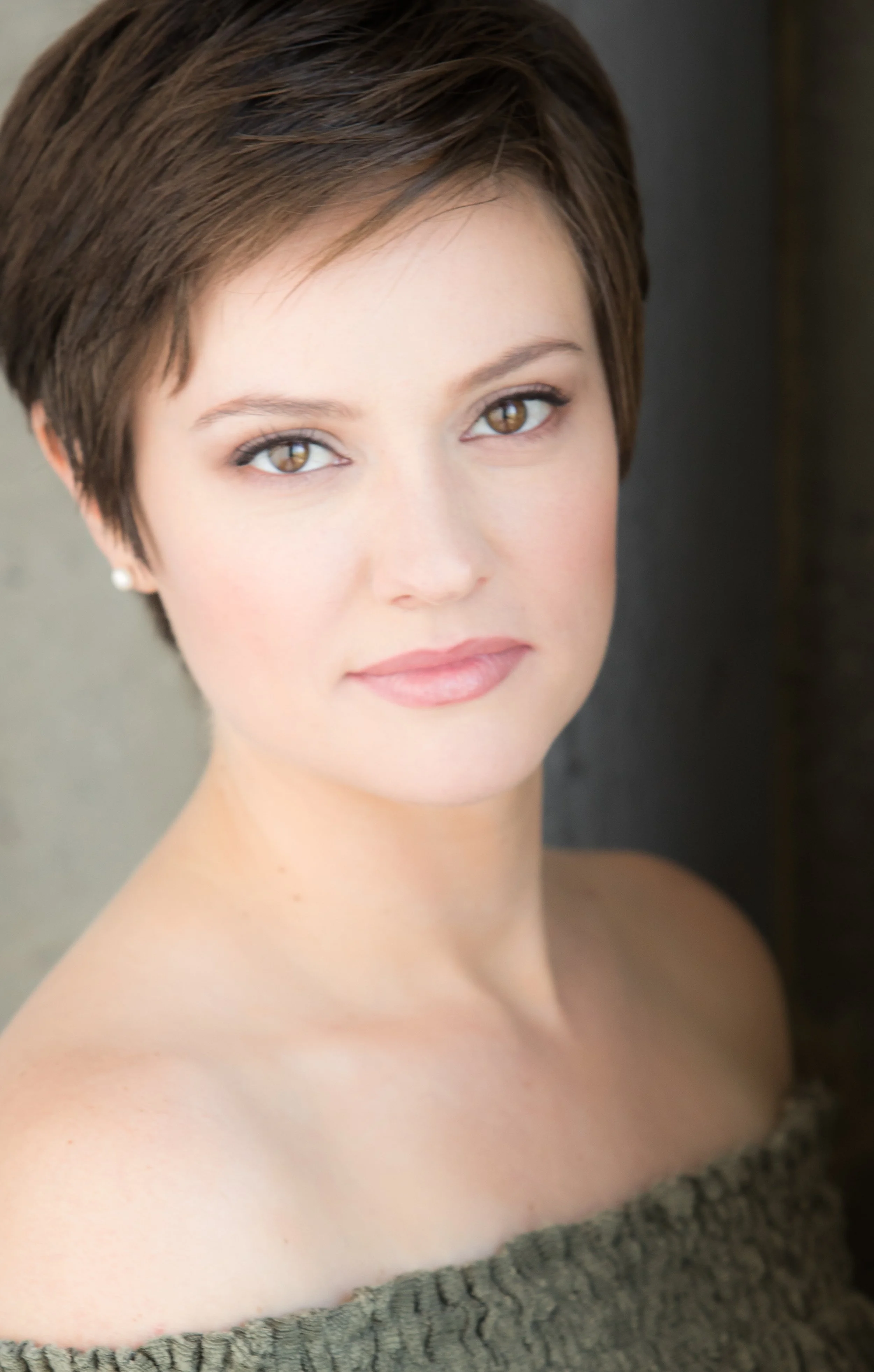 A woman with short brown hair, light skin, and brown eyes, wearing a gray off-the-shoulder sweater.