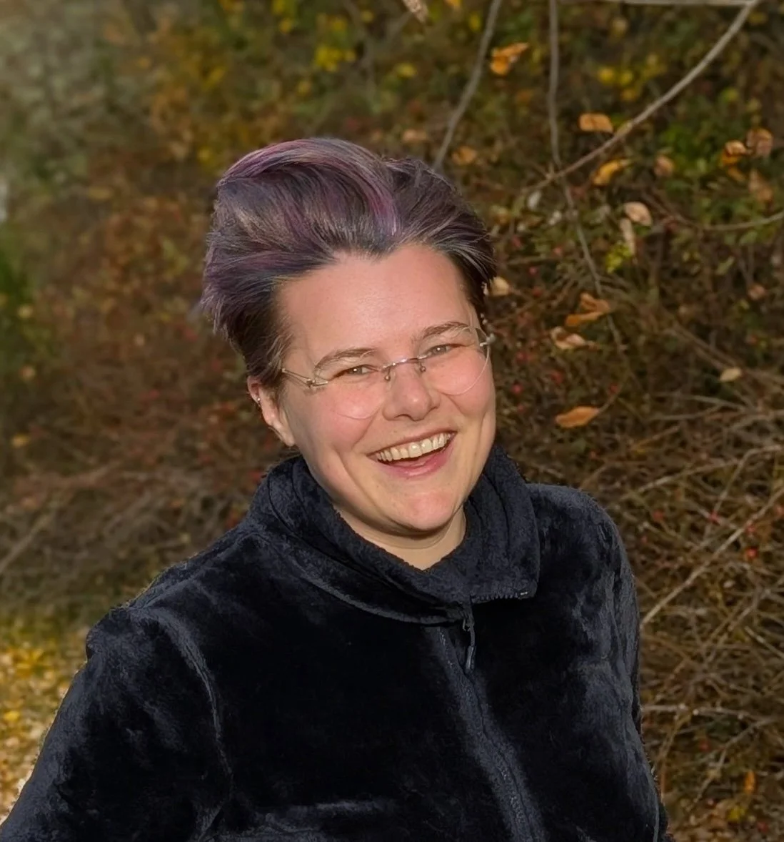 A smiling woman with short, dark hair with purple highlights, wearing glasses and a black jacket, standing outdoors with autumn foliage in the background.