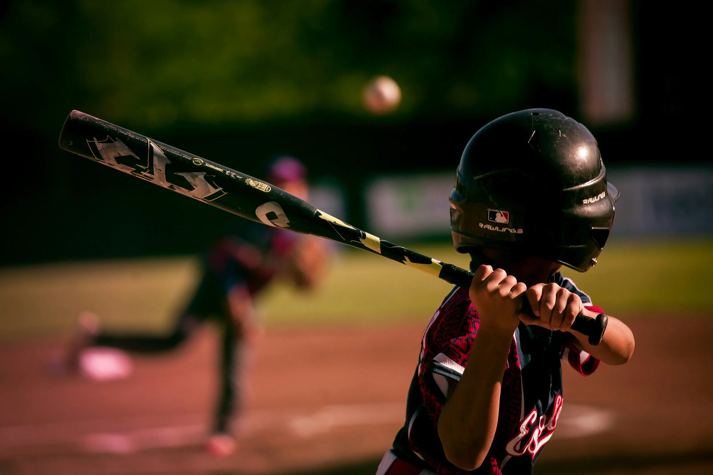 A boy wears a black baseball helmet and holds the bat ready to swing. The baseball is almost to him.