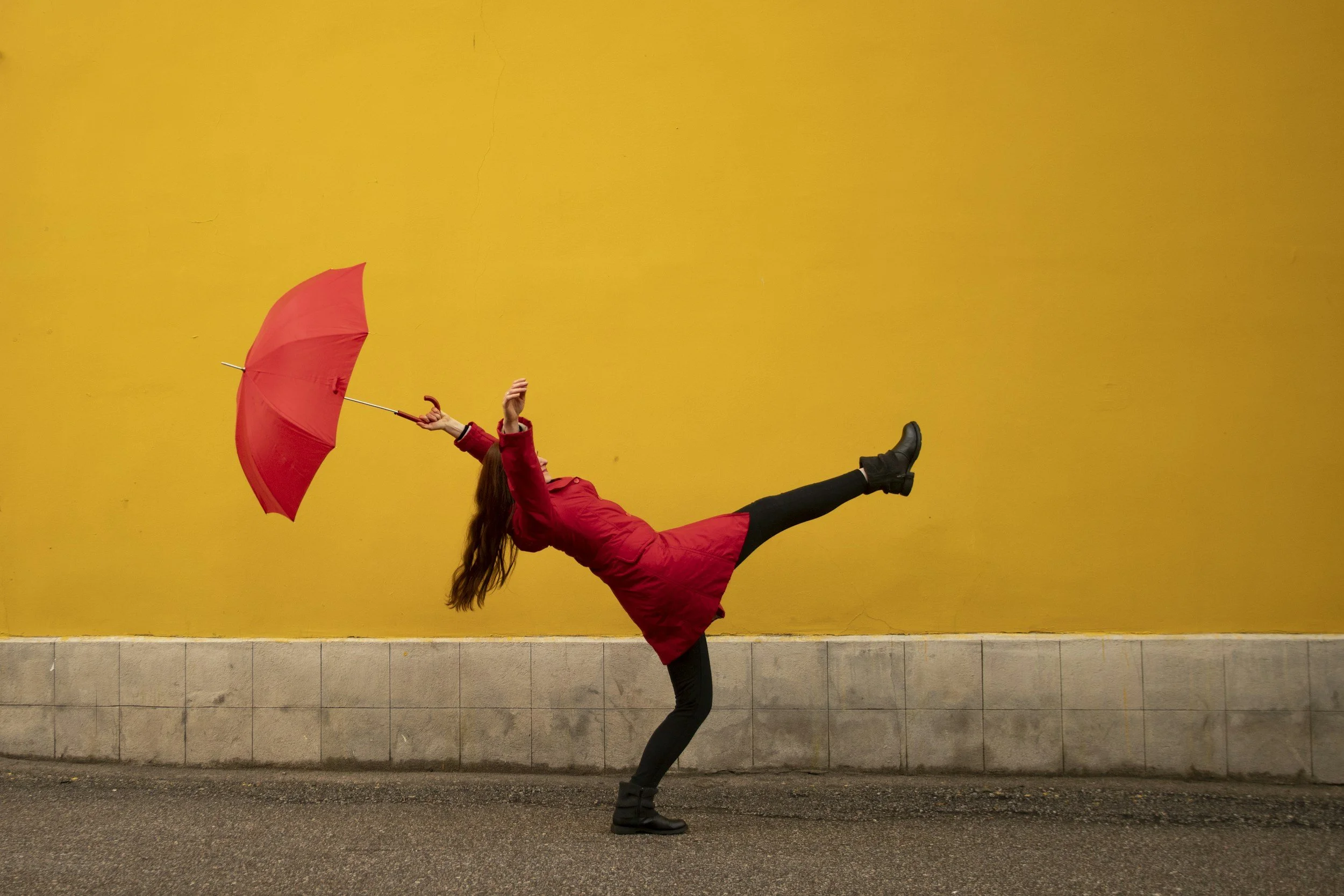 A woman in a red jacket and a red umbrella is standing on one foot in front of a yellow wall.