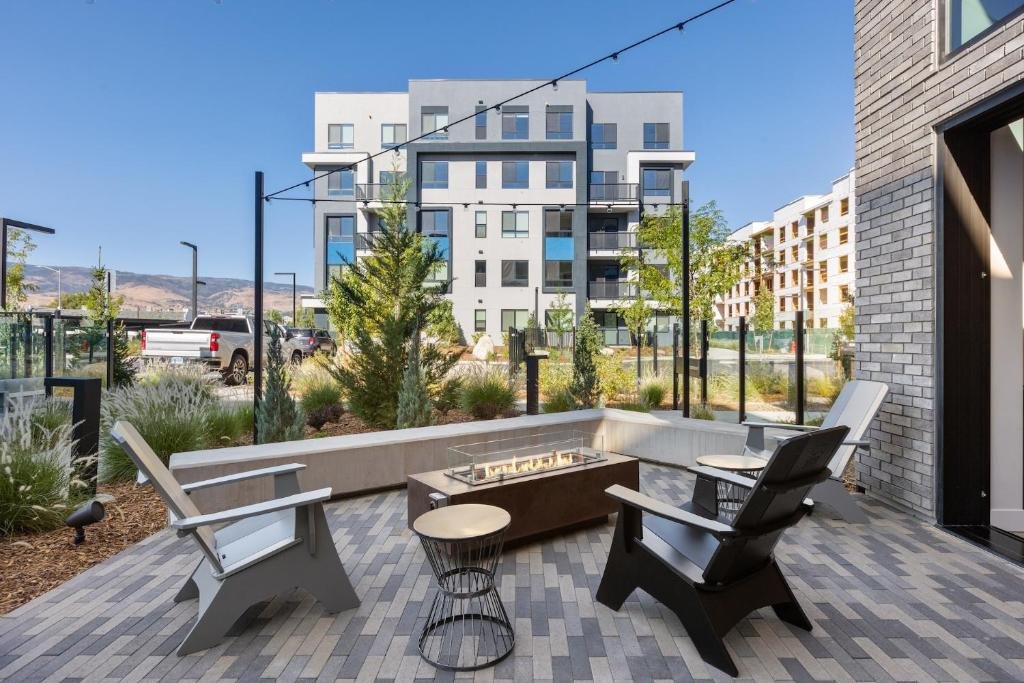 Outdoor patio with modern chairs and a built-in rectangular fire pit, surrounded by landscaping and glass fencing, with apartment buildings and mountains in the background.